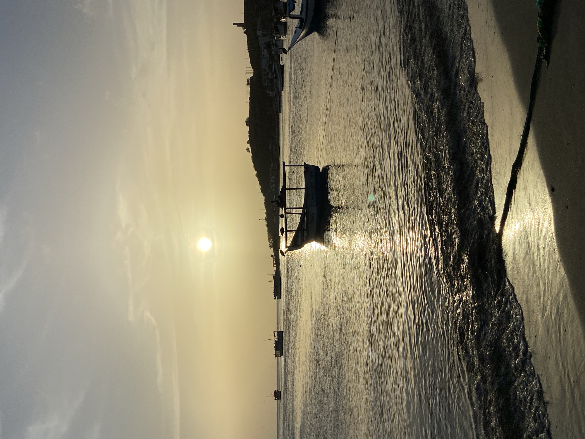The image depicts a serene sunset over the bay of Juan Griego, Venezuela. The sky is a soft gradient of pale yellow and grey, with the sun a bright, diffused orb near the center. The water is calm, reflecting the warm light of the setting sun, creating a shimmering path across its surface. Several small fishing boats are anchored in the bay, their silhouettes dark against the luminous water. In the foreground, a gentle wave laps onto a sandy beach, where a rope lies partially buried in the sand. The distant shoreline is characterized by a low, tree-covered hill dotted with buildings, including what appears to be a lighthouse or tower. The overall atmosphere is one of tranquility and natural beauty.
