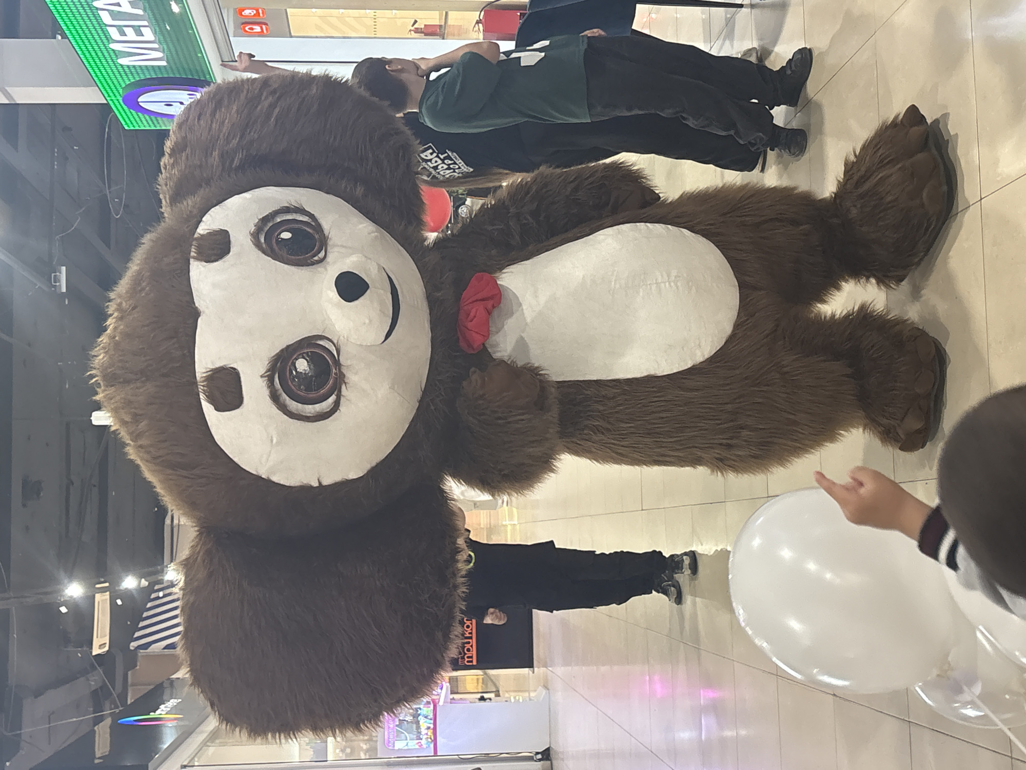 A mascot costume, likely a depiction of Cheburashka, is the primary subject of the image, appearing in a shopping mall. The costume is brown and white, with large ears and a red bow. Several people are in the background, including one person lying on a bench or ledge, and another walking. A child is holding white balloons in the foreground, with their hand partially visible. The mall features signs in Russian, including "MEGA," and various storefronts. The location is presumably Tri Potoka, Russia. The floor is tiled and the overall scene appears to be a public event or promotional activity within the shopping mall.