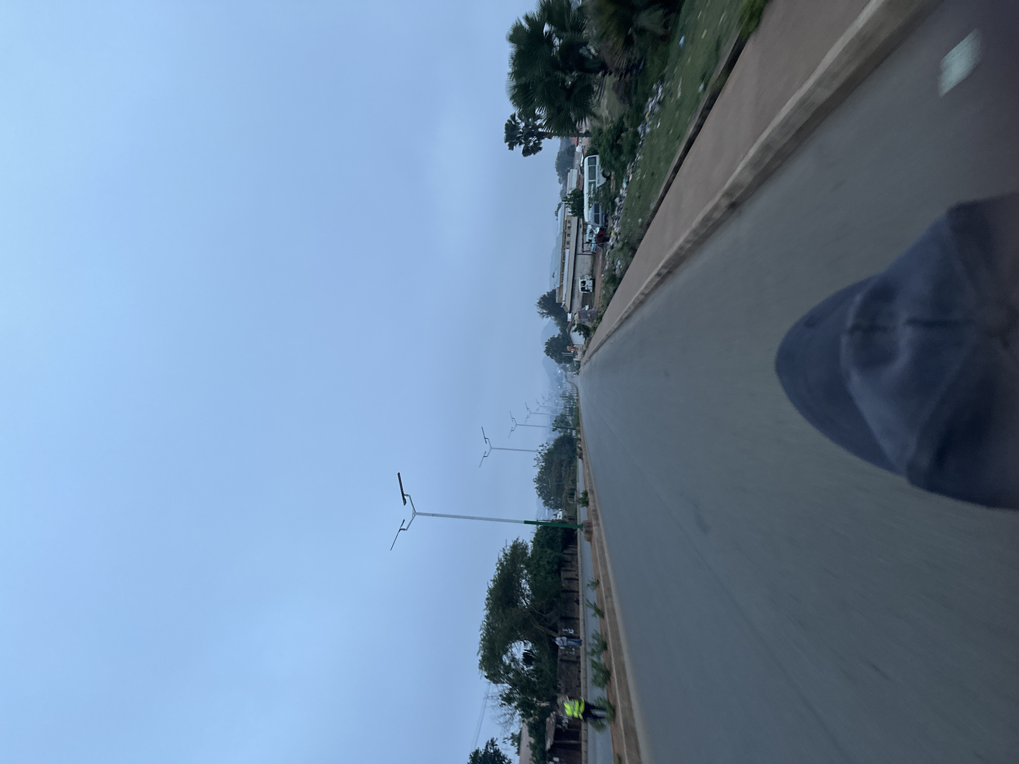The image depicts a street scene in Bukuru, Nigeria, under a cloudy sky. A paved road stretches into the distance, flanked by a curb and a roadside area with trees, vegetation, and occasional streetlights. Buildings and vehicles can be seen in the background. The foreground includes a partial view of a person.