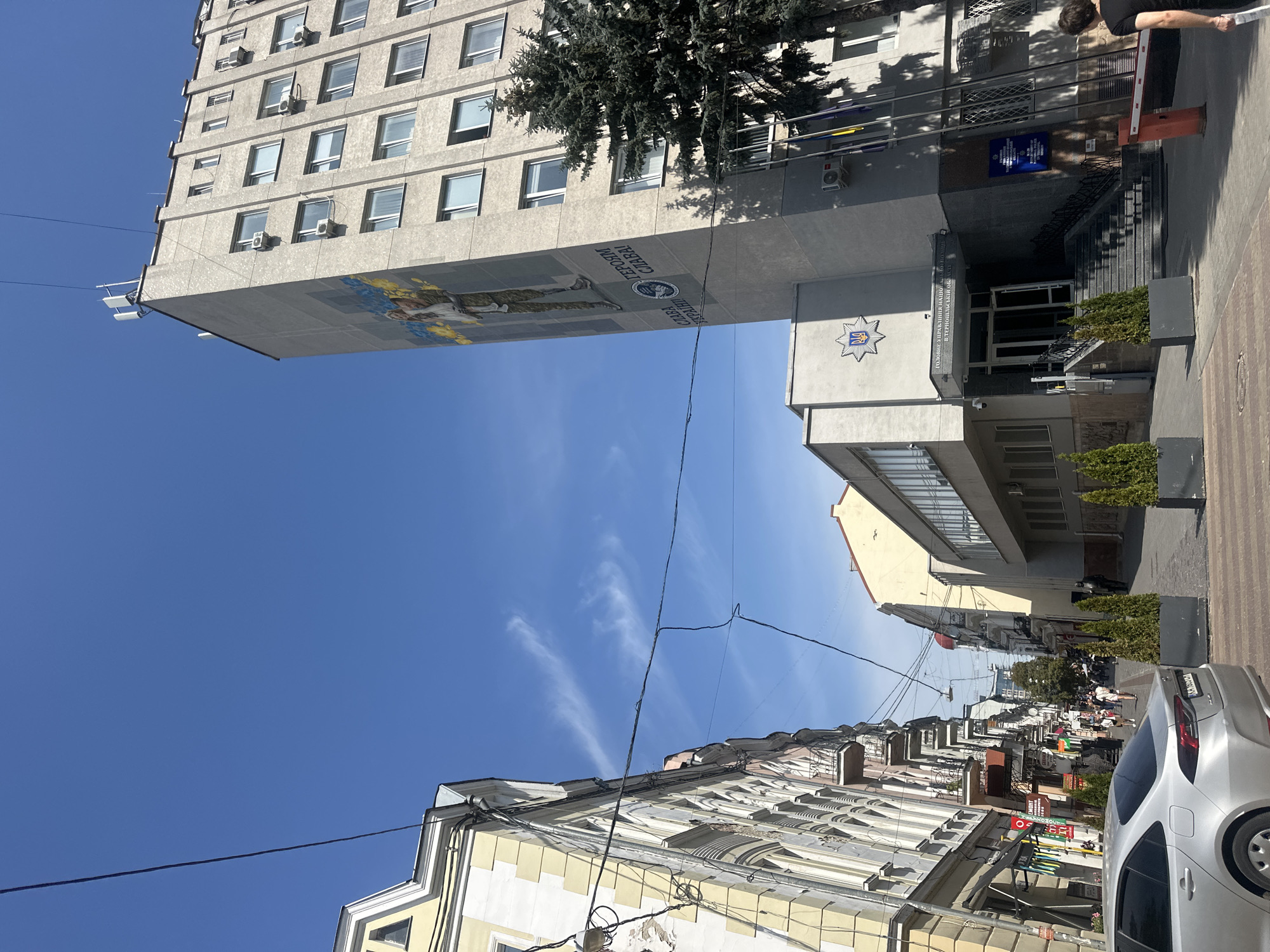 The image captures a street scene in Ternopil, Ukraine, on a clear, sunny day with a bright blue sky. The focal point of the scene is a tall, modern, concrete building with a large mural on its side. The mural depicts a soldier, possibly a tribute to Ukrainian military personnel, with the Ukrainian flag colors prominently featured. The building has numerous windows, and some appear to be open, with air conditioning units visible. The mural includes Ukrainian text that translates to "Glory to Ukraine! Glory to the heroes!"
In front of this building, a smaller, lower-rise structure houses what appears to be a police station, indicated by a blue sign with Ukrainian text and a police emblem. The entrance to the police station is flanked by steps and planters with evergreen trees. Several flags, including the Ukrainian flag, are displayed on poles near the entrance.
To the left of the police station and mural building, an older, ornate building with a light yellow facade and white trim lines the street. This building also has many windows and some architectural details like balconies and decorative elements. Further down the street, the view opens up to a row of buildings with a mix of architectural styles, suggesting a typical urban environment.
A silver sedan is parked on the street in the foreground, facing away from the viewer. A pedestrian crossing is visible in front of the police station. In the distance, a busy street with shops and pedestrians can be seen. A few people are walking on the sidewalk and crossing the street. The overall impression is a snapshot of daily life in a Ukrainian city, with a prominent display of national pride and support for its military.