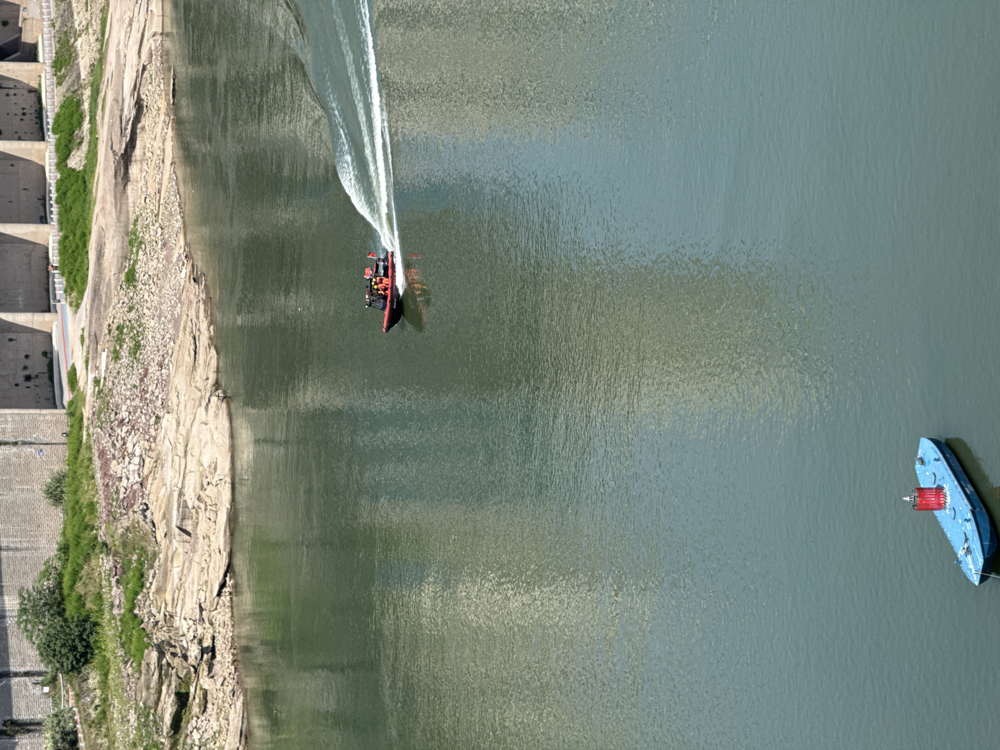 This high-angle, overhead photograph captures a wide expanse of greenish-blue water, likely a river or reservoir, bordered by a rocky shoreline and a massive concrete dam-like structure. The scene unfolds under bright daytime conditions, indicated by the strong reflections and ripples on the water's surface.
In the center of the frame, a red and black rigid inflatable boat (RIB) or patrol boat is actively traversing the water, creating a prominent white wake in its path. Several individuals, visibly wearing orange life vests, are on board, suggesting a crew or passengers engaged in navigation or patrol. Towards the right side, a stationary blue boat, possibly a work platform or barge, is anchored or moored, featuring a distinct red cylindrical object on its deck, which may serve as a light or marker.
The left side of the image reveals a rugged, rocky bank adorned with sparse green vegetation, leading up to a monumental concrete structure. This structure, characterized by multiple buttresses or sections, strongly resembles a dam, flood barrier, or significant river infrastructure. A walkway or road runs along its top, where a lone human figure can be faintly observed, providing a sense of scale.
The activity primarily involves the movement of the red and black boat, while the blue boat remains static. The setting, within Yuzhong District, China, aligns with the confluence of major rivers like the Yangtze and Jialing in Chongqing, known for their extensive river infrastructure. No discernible text is visible in the media file.