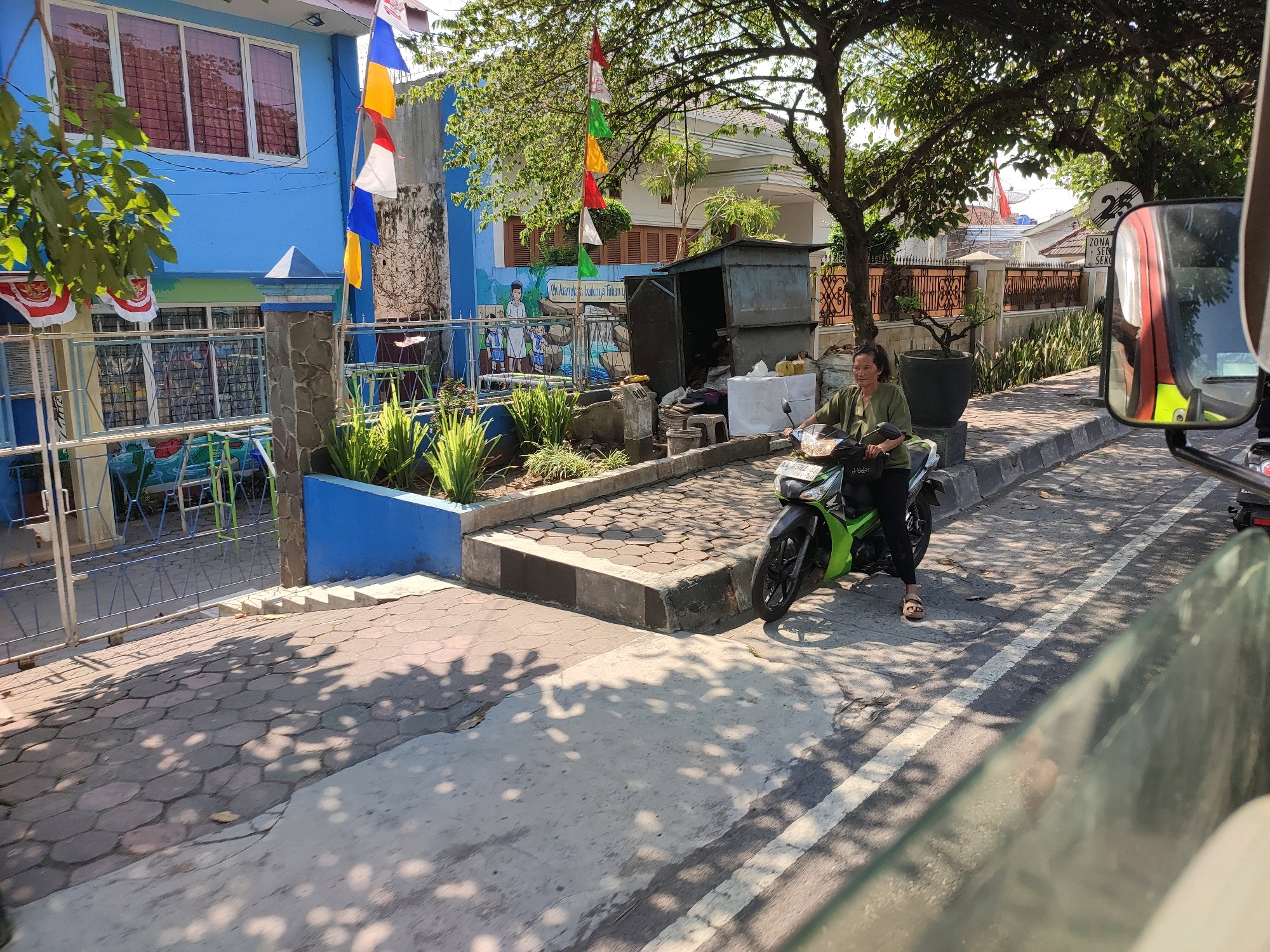 A woman is riding a green motorcycle on a street in Magelang, Indonesia, in front of a blue building with flags. The building appears to be a school, with children painted on the exterior wall and playground equipment visible through a fence. The woman is wearing a green tunic and black pants, with sandals. She is looking to her left. The street is paved with concrete and bordered by a sidewalk. Trees with dappled sunlight cast shadows on the ground. A street sign is visible in the background. The weather is sunny.