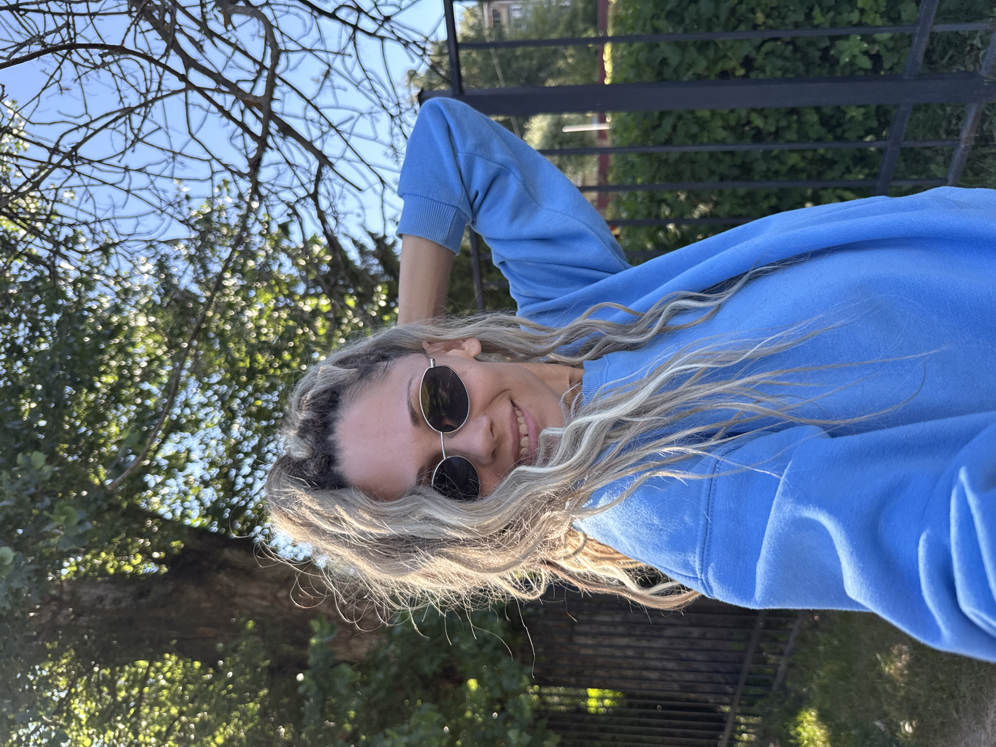 A woman with blonde hair pulled into a ponytail, wearing sunglasses and a blue sweatshirt, stands in front of a metal fence with greenery behind her. Sunlight filters through the branches of a large tree, casting dappled light on the scene. The sky is clear and blue. The location is identified as Rusanovo, Russia.