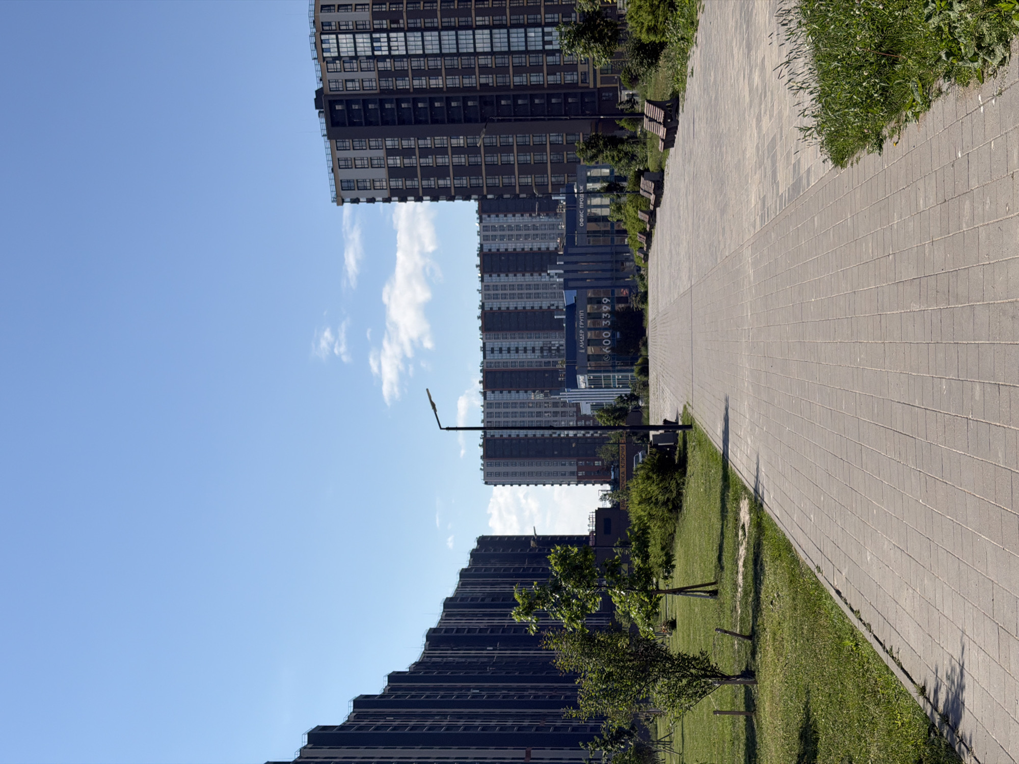 The image depicts a wide paved pathway in a park-like setting in Saint Petersburg, Russia. On the left and right sides of the pathway are grassy areas with young trees and bushes. In the background, a series of modern, multi-story residential buildings are visible. The buildings are dark in color with numerous windows. A light blue sky with a few scattered clouds is overhead. Several park benches are placed along the right side of the pathway. A streetlamp is visible in the middle ground of the pathway. There are no people or animals visible in the image. The scene appears to be a well-maintained urban public space on a clear day.
