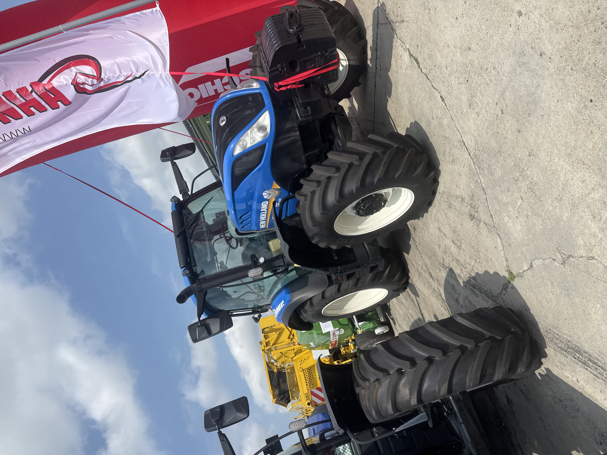 This media file presents an outdoor scene featuring agricultural machinery, likely at an exhibition or dealership. The primary subject is a large, modern blue New Holland T6.145 tractor, positioned on concrete ground under a bright, partly cloudy sky.
The tractor is prominently displayed, featuring large, rugged tires with white rims and black counterweights at the front. Its body is a vibrant blue with black accents around the grill and headlights. Behind the tractor, parts of other machinery are visible, including a bright yellow piece of equipment with the number "380" clearly marked, and a green machine partially visible further back.
In the upper left portion of the frame, a white flag with a distinct red and black logo or partial text (possibly "HAA") is visible, tethered to a red banner. The banner itself bears the partially visible text "SCHICHO". This flag and banner arrangement suggests an outdoor event or display, such as a trade fair or open day.
The time of day is clearly sunny, with bright daylight casting noticeable shadows on the concrete ground. The sky is blue with scattered white clouds, indicating pleasant weather. The concrete surface shows some cracks and irregularities. The image's original orientation places the tractor vertically on its side, but the scene is best interpreted as a horizontal view of machinery on display in an open area. The context suggests the location is Stara Zagora, Bulgaria.