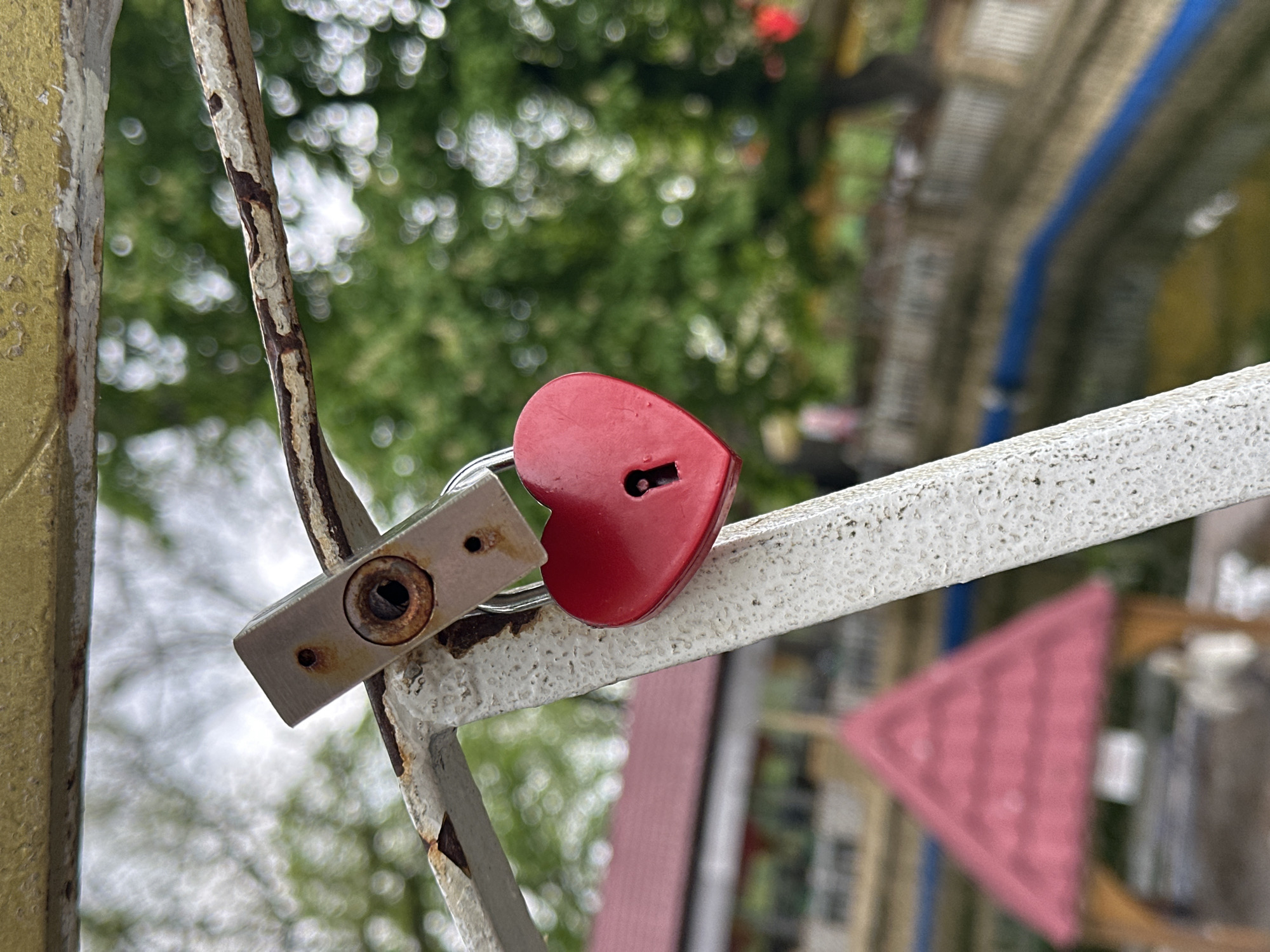 The image shows a red heart-shaped padlock attached to a white metal railing. Next to the red padlock is a silver, rectangular padlock, also attached to the railing. Both padlocks appear to be old, with visible signs of wear and rust. The background is blurred, but greenery and architectural structures can be discerned, indicating an outdoor location. Based on the location provided, this could be the “Tree of Love” site on a bridge in Stavropol, Russia, where couples often place padlocks to symbolize their enduring love.