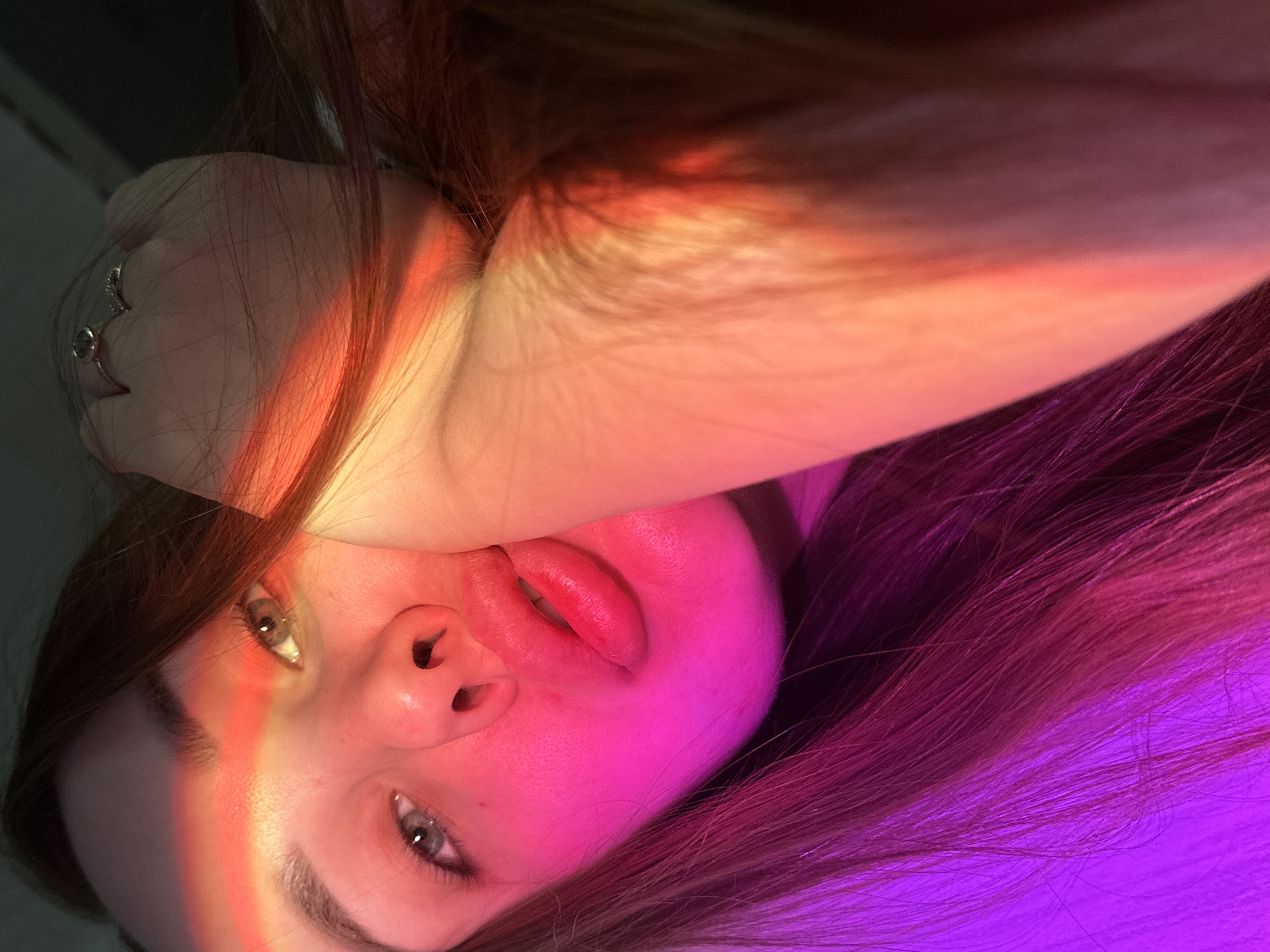 A close-up, tilted selfie-style photograph shows a woman's face with her right arm raised and hand near her forehead. She has light skin, visible gray-green eyes, and prominent eyebrows. Her lips are full and reddish. A beam of multicolored light, from red to yellow, shines across her face and hand, while the lower part of her face and hair are illuminated with purple light. She has brown hair that falls loosely around her face. A ring is visible on one of the fingers of her right hand. The background appears to be a dark, indistinct surface, possibly a wall or ceiling. It is not possible to determine the exact location of the photo, specifically within the city of Chaikovsky, Russia, without further information or context.