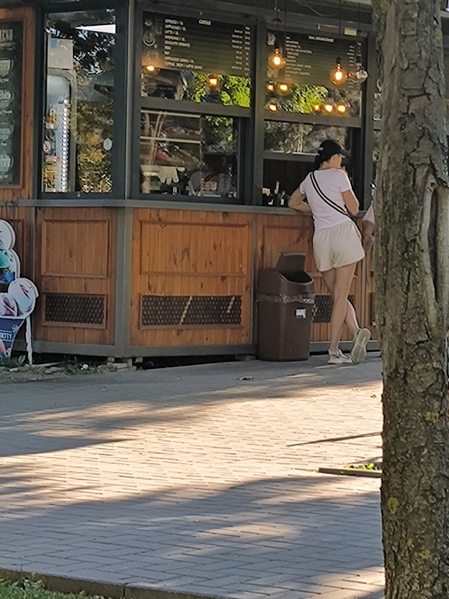 The image depicts a street-level view of a wooden kiosk, likely a food or beverage vendor, situated outdoors. A woman wearing a light-colored t-shirt, white shorts, and a black cap is standing at the counter of the kiosk, facing away from the camera and engaging with someone inside. She has a black and white strap across her chest. Another person is partially visible behind her. A brown trash bin stands to the left of the woman. The kiosk features a menu board with text and illuminated bulbs visible through a glass window. The foreground consists of a paved walkway bathed in dappled sunlight, with shadows cast by trees. The background shows greenery, suggesting a park or tree-lined street. The overall scene is consistent with an outdoor commercial area in Chișinău, Moldova, likely a public space like a park or pedestrian zone where such vendors are common.
