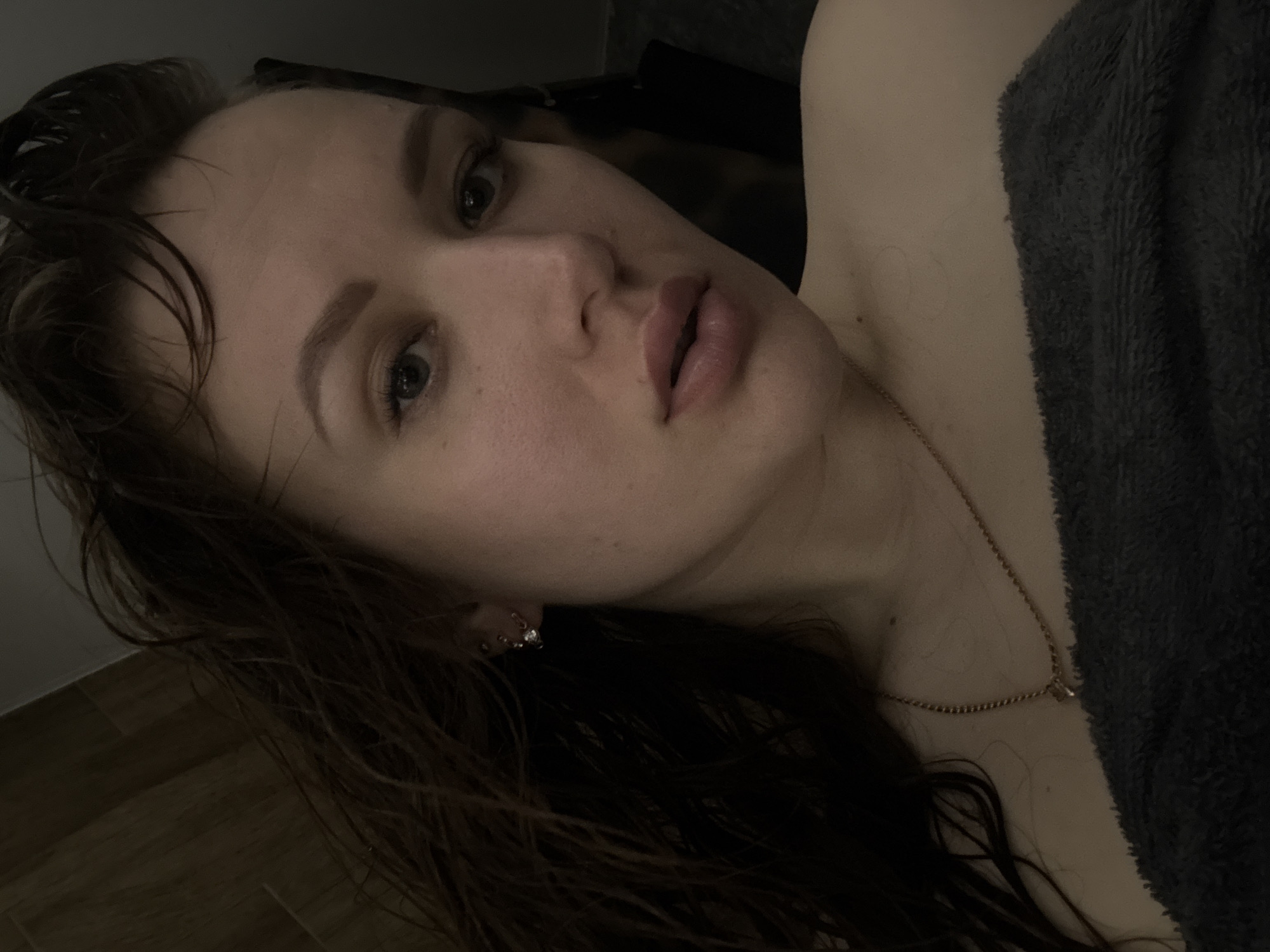A close-up, angled portrait shows a young, light-skinned woman with damp, dark brown hair and light-colored eyes. She is positioned to the left of the frame with her head tilted to her left. She has full lips and is wearing a small stud earring. A fine gold chain necklace is visible against her skin. To the right, a gray, textured material, possibly a towel or blanket, frames the edge of the image. The background is dimly lit, suggesting an indoor setting, with hints of wooden flooring visible in the lower left. The scene suggests a personal or private space, possibly a bathroom or bedroom. The photograph does not provide any location information for Chaikovsky, Russia.