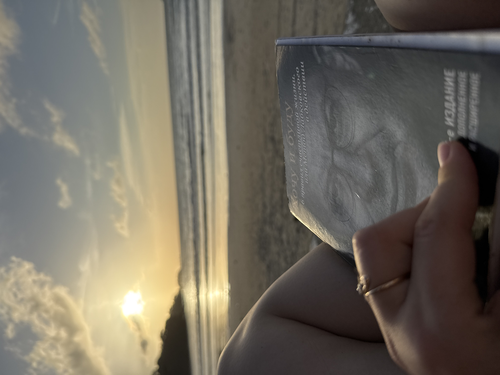 The image depicts a close-up view of a person holding a book while sitting on a beach at sunset. The book's cover features a portrait of a man wearing glasses, and some text in Russian. The text appears to be a title and possibly some subtitle information, possibly related to psychology or self-help. The person's hand, adorned with a ring, is visible holding the book. The background shows a beach with sand, the ocean with gentle waves, and a sky illuminated by the setting sun. The overall scene suggests a moment of relaxation and reading in a scenic, coastal environment. The location is specified as the city of Patong, Thailand.