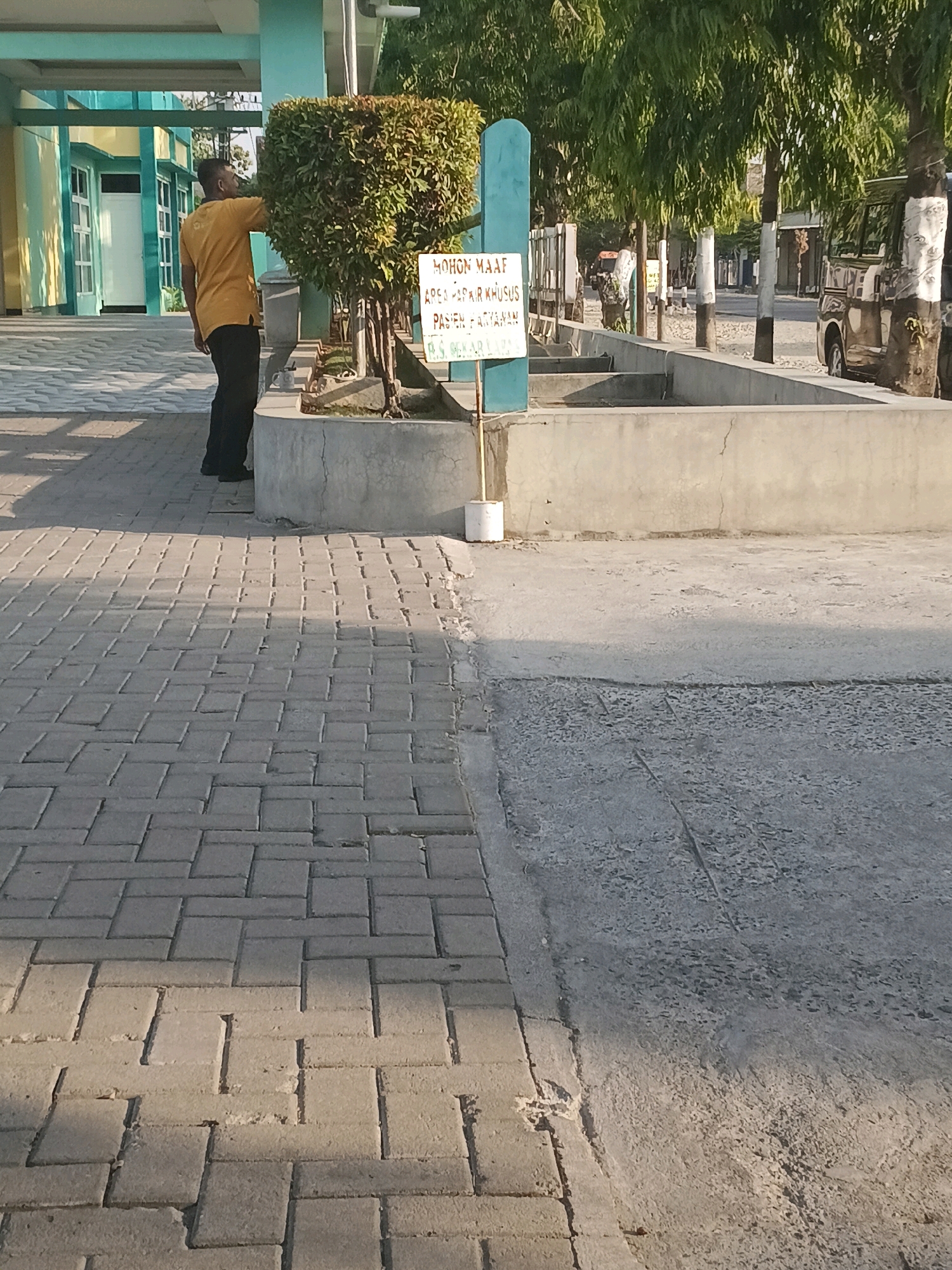 A person in a yellow t-shirt and black pants stands near a trimmed hedge and a sign in front of a building in Purwodadi, Indonesia. The sign, written in Indonesian, reads "MOHON MAAF AREA PARKIR KHUSUS PASIEN & TAMU" which translates to "SORRY, PARKING AREA IS EXCLUSIVELY FOR PATIENTS & GUESTS". The ground is paved with interlocking grey bricks in the foreground, and a concrete walkway with scattered debris is visible further back. Trees with green foliage line the background. A dark-colored van is parked to the right. The building behind the man has light green and yellow walls with white-framed windows and doors. The overall scene depicts a sunny outdoor environment.