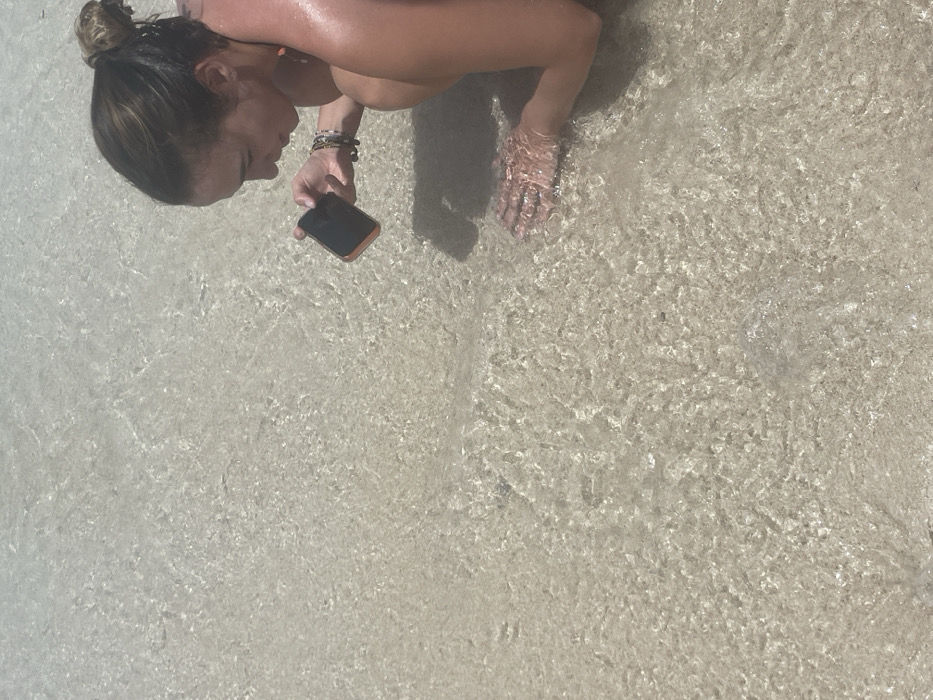 An adult female, with tanned skin and dark hair tied into a bun, is observed squatting in shallow, clear water over a light sandy bottom. This scene is consistent with a coastal area in the city of Zanzibar, Tanzania. She wears what appears to be an orange-strapped swimsuit and multiple bracelets on her left wrist. Her head is tilted downwards, eyes focused intently on the water directly in front of her. She holds a black smartphone, encased in an orange protective cover, in her right hand, pointing it towards the water's surface. Her left hand is submerged in the clear water, with fingers slightly spread. The water surface exhibits distinct ripples caused by light refraction from an overhead light source, likely the sun, which illuminates the scene brightly.