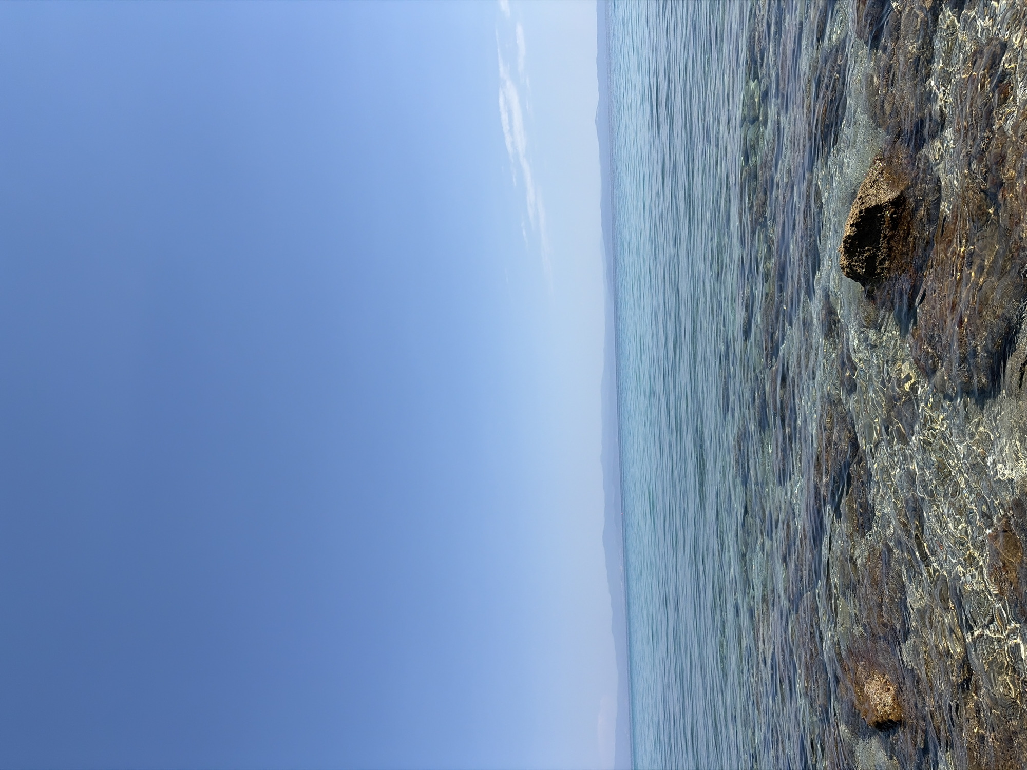 The image displays a serene seascape from a clear, shallow body of water looking towards a distant, hazy shoreline under a bright, cloudless blue sky. The foreground consists of a rocky seabed visible through the translucent water. Numerous stones, varying in size and color from dark brown to lighter reddish hues, are scattered across the bottom. The water itself is calm, with gentle ripples reflecting the sunlight and creating a shimmering effect. A few larger, distinct rocks are partially submerged and break the surface.
In the middle ground, the water deepens slightly, transitioning to a deeper, more uniform turquoise color. The surface of this section of the sea is marked by subtle wave patterns.
The background features a long, low-lying mountain range that stretches across the horizon, appearing muted and distant due to atmospheric haze. The sky above is an expansive and uninterrupted expanse of vibrant blue, with a single, thin wisp of a cloud visible near the horizon on the right side.
This scene is characteristic of the coastal areas within the Kassandra Municipal Unit in Greece, specifically the Halkidiki peninsula, known for its clear waters and picturesque coastlines. The image captures a quiet moment, with no people or vessels visible, emphasizing the natural beauty of the environment.