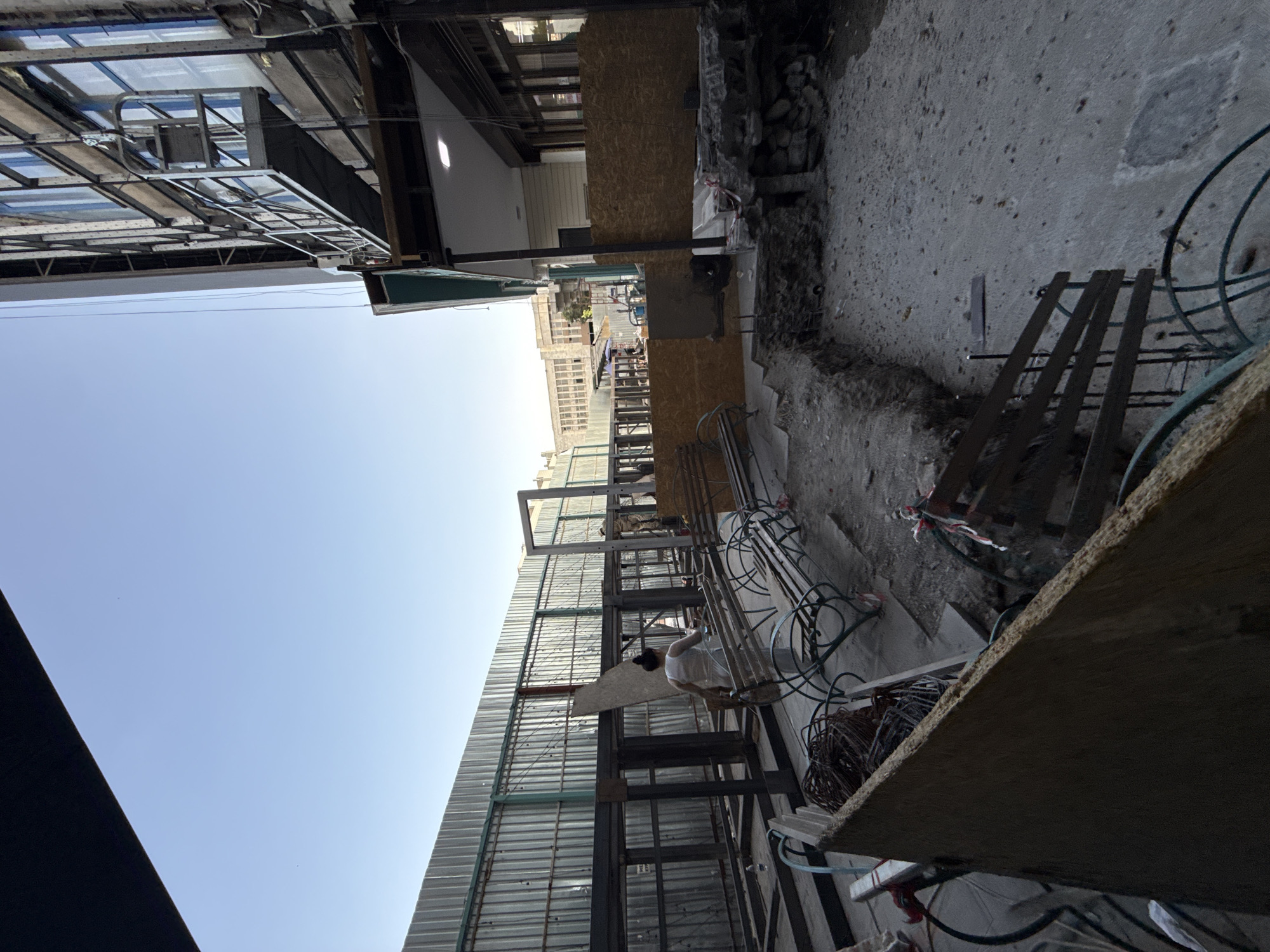 A wide-angle, eye-level shot captures an outdoor construction site. The scene is predominantly concrete and metal, with a clear blue sky overhead. In the foreground, a large sheet of plywood is angled from the bottom left. Behind it, construction materials like wire mesh and rebar are visible.
To the left, a metal fence stretches into the distance, part of a larger structure under construction. In the middle ground, a woman in a white outfit walks away from the camera, carrying a piece of material. Several empty metal benches with decorative scrollwork are placed in front of the unfinished structure.
The right side of the image shows the side of a building with multiple windows and scaffolding. A suspended platform is attached to the building's exterior. In the foreground on the right, a section of dug-out earth reveals stone and concrete foundations, with exposed rebar and wiring. The ground is rough concrete and dirt. The overall impression is one of ongoing urban development and renovation. The location is identified as Bishkek City, Kyrgyzstan.