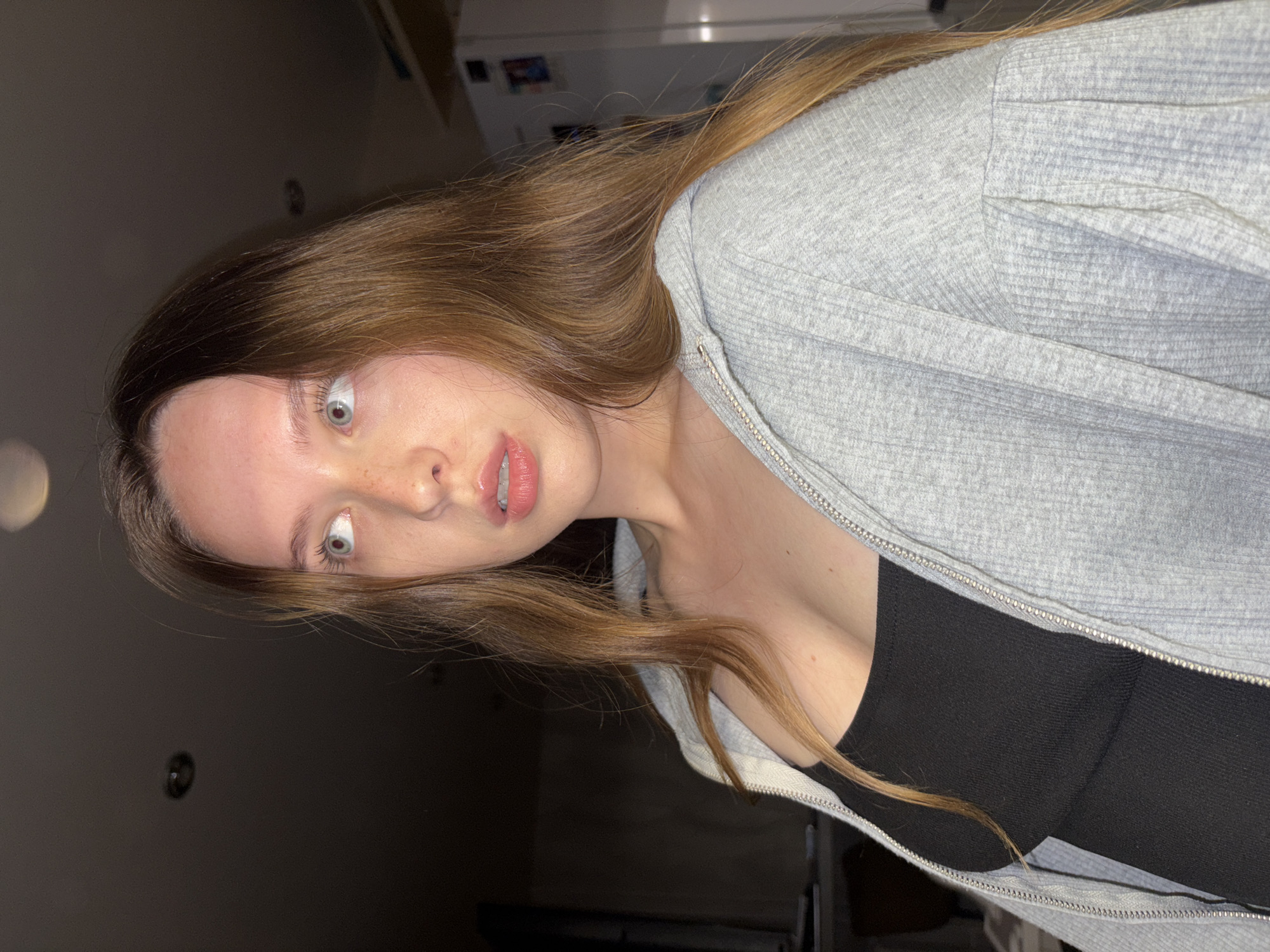 The image shows a medium close-up of a young woman with long, auburn hair. She is looking to her left, with her mouth slightly open. She is wearing a black tank top and a grey zippered hoodie. The background is dark and out of focus, with some indistinct objects that appear to be in an indoor setting. There are ceiling lights visible in the upper portion of the image. The scene appears to be an indoor residential environment, possibly a home, in the city of Chaikovsky, Russia.
