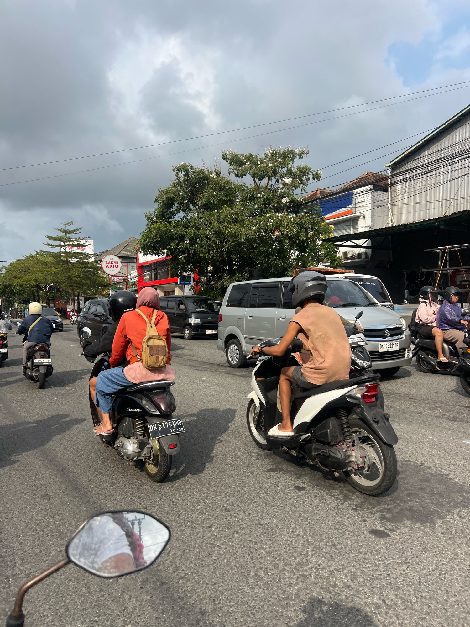 The image captures a street scene in Denpasar, Indonesia, likely during daylight hours under a cloudy sky. The foreground shows the handlebar and mirror of a motorcycle, reflecting a person's legs and part of a red garment. The mid-ground features several motorcycles and vehicles in motion on an asphalt road.
Prominently, two individuals are riding a black scooter. The rider in front, wearing a light brown sleeveless shirt and dark shorts, is facing away from the camera. The passenger behind, wearing an orange jacket and a pink hijab, also faces away, with a beige backpack slung over one shoulder. Their scooter has a license plate that reads "DK 5176 UAD".
To the left, another person on a black scooter wears a blue jacket and a white helmet, also moving away. Further back, a silver minivan and other vehicles are visible, indicating moderate traffic. On the right side of the road, buildings with corrugated metal roofs and signage line the street. One sign displays "BAKMI AKIU" and "HALAL". Trees with green foliage are interspersed along the roadside. The overall atmosphere suggests a busy urban environment.