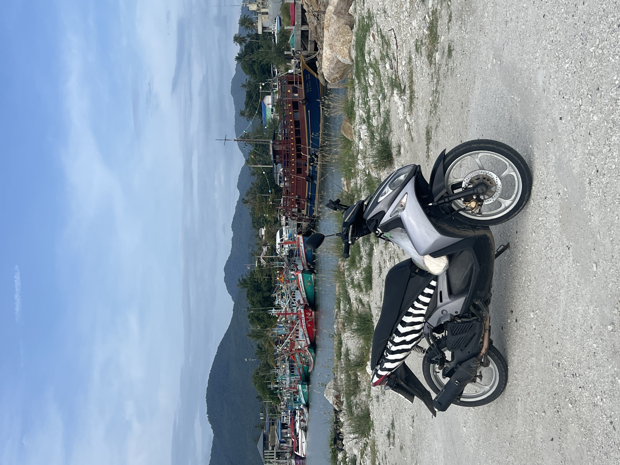 This image shows a scenic harbor on Ko Pha-ngan District, Thailand. The foreground is dominated by a silver and black scooter parked on a gravelly path. The scooter has a black seat with a distinctive black and white striped cover. Beyond the scooter, a calm body of water is filled with numerous fishing boats of various sizes, their colorful hulls and intricate rigging creating a vibrant scene. In the background, lush green mountains rise up, their slopes dotted with palm trees. A large, multi-decked wooden boat with a dark blue hull and red accents is prominently moored, adding a touch of historical charm to the harbor. The sky is a soft blue with scattered white clouds, suggesting a pleasant day. The overall impression is one of a peaceful, working harbor in a tropical paradise. There is some visible text on the side of the large wooden boat, but it is too small to be legible in this image.
