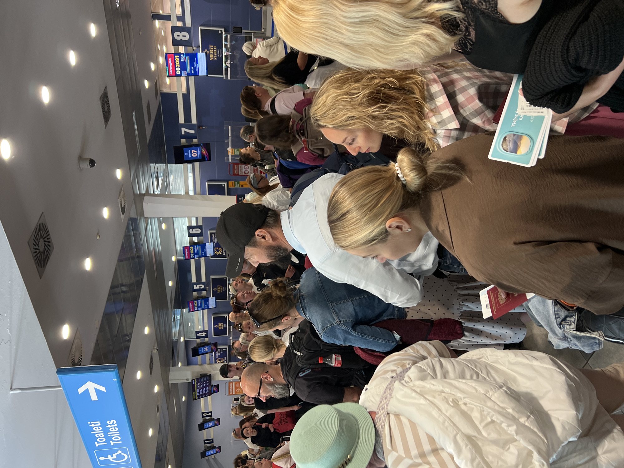 The image depicts a busy airport departure hall, likely at Tivat Airport in Mrčevac, Montenegro, given the context of a flight. A large crowd of diverse individuals stands in a queue, with many looking towards the check-in counters. Several digital displays are visible, indicating gate numbers and flight information, including a Norwegian flight DS2091 to Riga at gate 8, and another to Helsinki at gate 5. A blue sign with white text and an arrow points towards "Toaleti Toilets" with a wheelchair accessibility icon. People are dressed in casual travel attire, carrying luggage and some are holding passports and boarding passes. The environment appears to be an indoor terminal with bright overhead lighting and a tiled floor. The overall atmosphere suggests a typical airport scene with passengers awaiting departure.