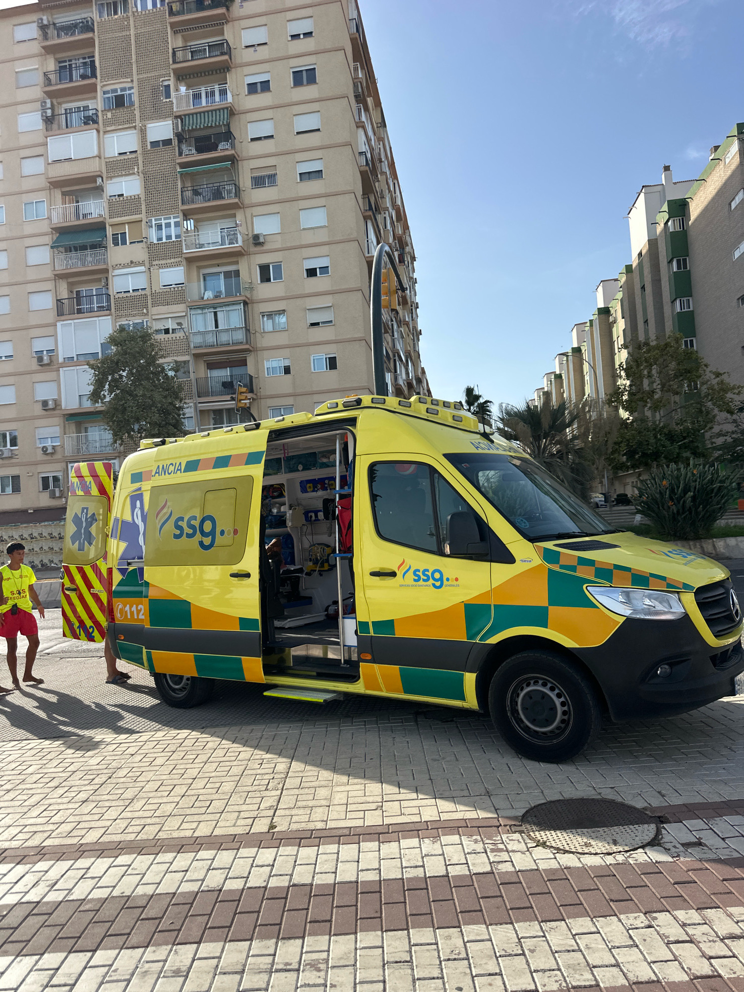 In Málaga, Spain, an ambulance is parked on a paved pedestrian walkway. The ambulance, predominantly yellow and green, has its rear and side doors open, revealing medical equipment inside. A young person wearing a yellow shirt and red shorts stands near the rear of the ambulance, facing away from the camera, with their left foot on the ambulance step. Another person's legs are visible near the open side door. In the background, tall apartment buildings line the street under a clear blue sky. The sunlight casts long shadows on the patterned pavement.