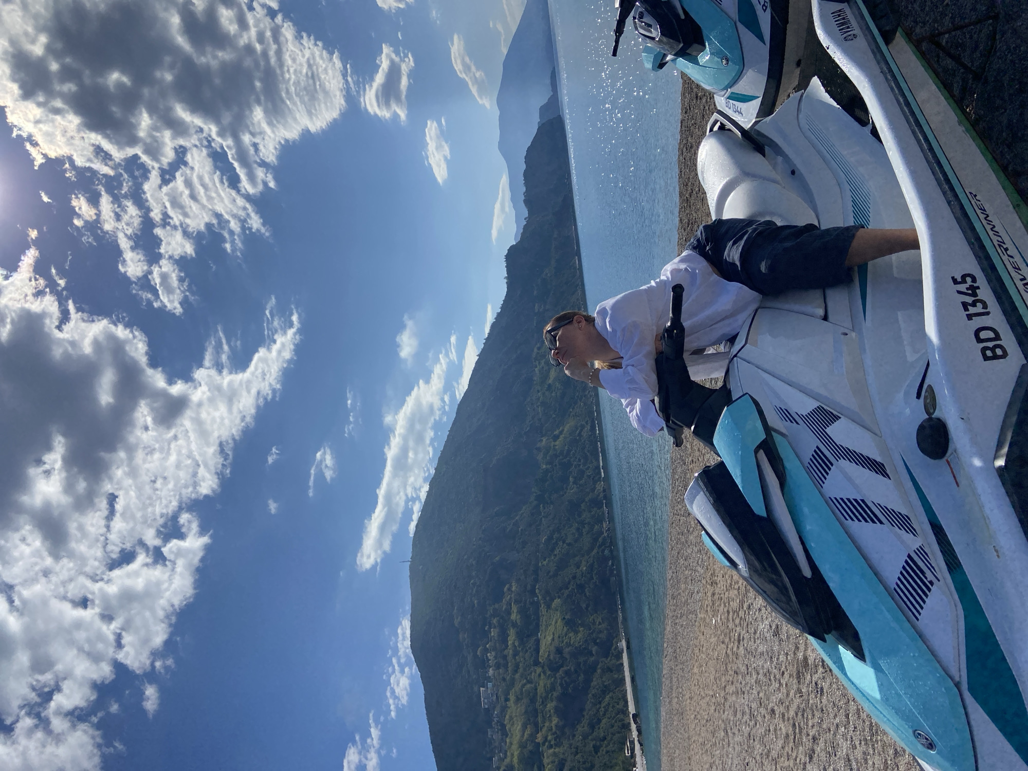 The image presents a daytime scene in Prijevor, Montenegro. The view includes a mountain range along a coastline where a person is seen lounging on a white and blue Yamaha WaveRunner personal watercraft. Another similar watercraft is positioned nearby. The water is sparkling, and the sky above displays a mix of clear blue and scattered white clouds. The person is wearing dark shorts and a white long-sleeved shirt, with sunglasses resting on their face.