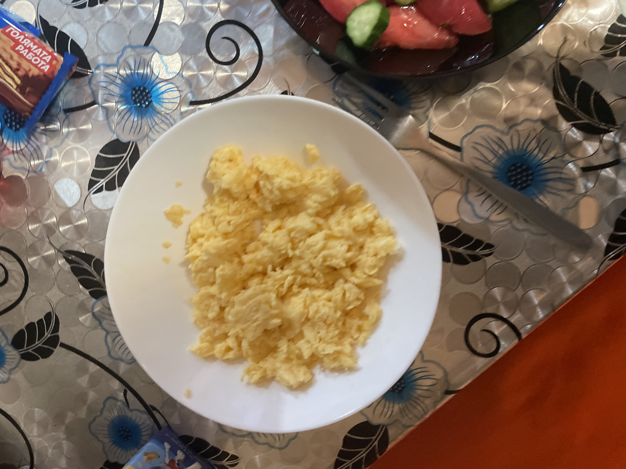 An overhead shot captures a simple meal set on a patterned tablecloth. The central focus is a white, round plate holding a generous portion of fluffy, yellow scrambled eggs. To the upper right, partially visible in a dark bowl, is a fresh salad consisting of diced red tomatoes and green cucumber slices. A silver fork rests between the scrambled eggs plate and the salad bowl.
In the upper left corner, a blue and red wrapper is visible. The text "ГОЛЯМАТА РАБОТА" (GOLYAMATA RABOTA), which translates to "The Big Job" or "The Great Work," is clearly printed on it in white Cyrillic letters, suggesting a snack bar or similar packaged food item.
The tablecloth features a distinctive design with metallic-looking concentric circles, interspersed with blue floral patterns and black leaf and swirl motifs. In the bottom right of the frame, an orange surface, possibly another part of the table or a different object, creates a contrasting border. The scene depicts a prepared meal, likely breakfast or a casual meal, laid out on a domestic table. No people are visible, and no specific time of day or emotional context can be discerned from the image alone.