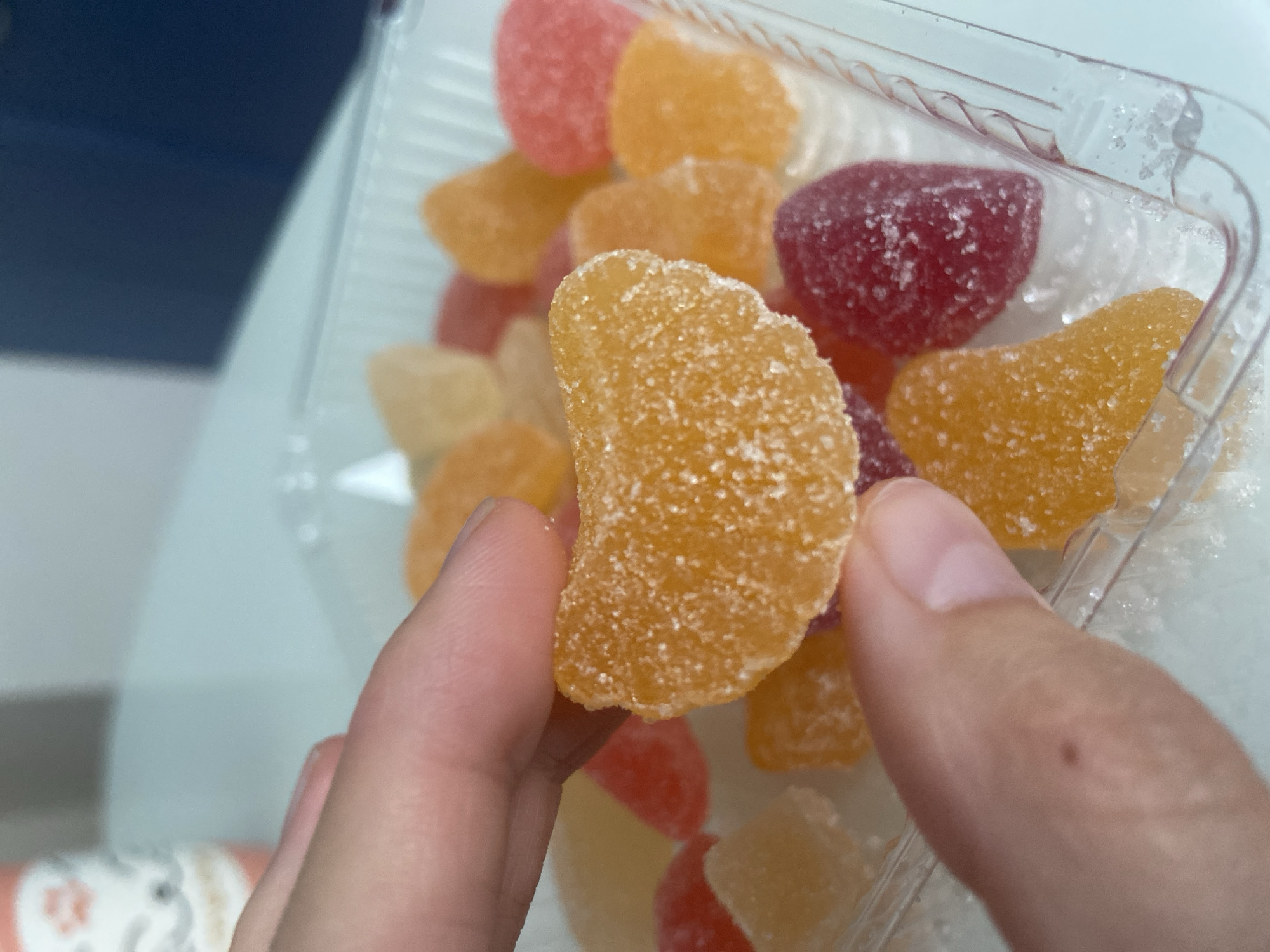 A close-up shot focuses on a hand holding an orange, crescent-shaped gummy candy coated in sugar. In the background, a clear plastic container holds an assortment of colorful gummy candies, including red, orange, and yellow pieces. The setting appears to be indoors, with a blurred background suggesting a table and possibly other objects. The lighting is bright and natural. The image does not provide any information related to the city of Balashikha, Russia.