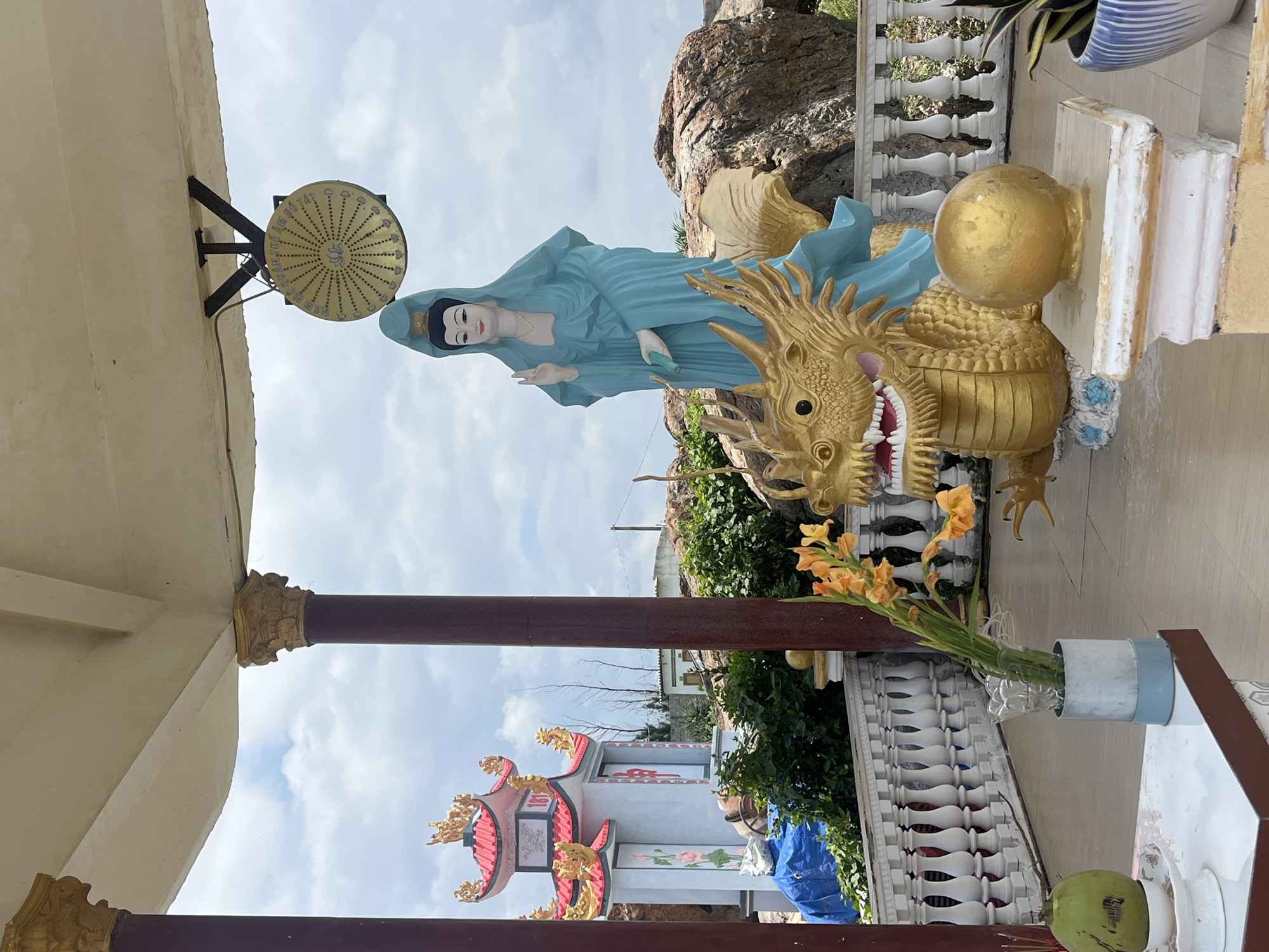 The image depicts an outdoor scene at a religious site in Mui Ne, Vietnam, under a cloudy sky. In the foreground, a white table holds a coconut, a white stand with incense, and a vase of orange flowers. To the right, a golden dragon statue is prominently displayed, with a serene statue of a woman in blue robes standing behind it. The dragon is intricately detailed, with visible scales, teeth, and claws. The woman's statue has a gentle expression and is depicted with her hand raised in a gesture of blessing. Behind these statues, a white balustrade lines the area, and a large rock formation can be seen. In the background, a small, ornate pagoda-style building with red and gold accents is visible. The overall atmosphere is one of reverence and tranquility, characteristic of a Buddhist or Taoist temple. The architecture and iconography suggest a location within Vietnam.