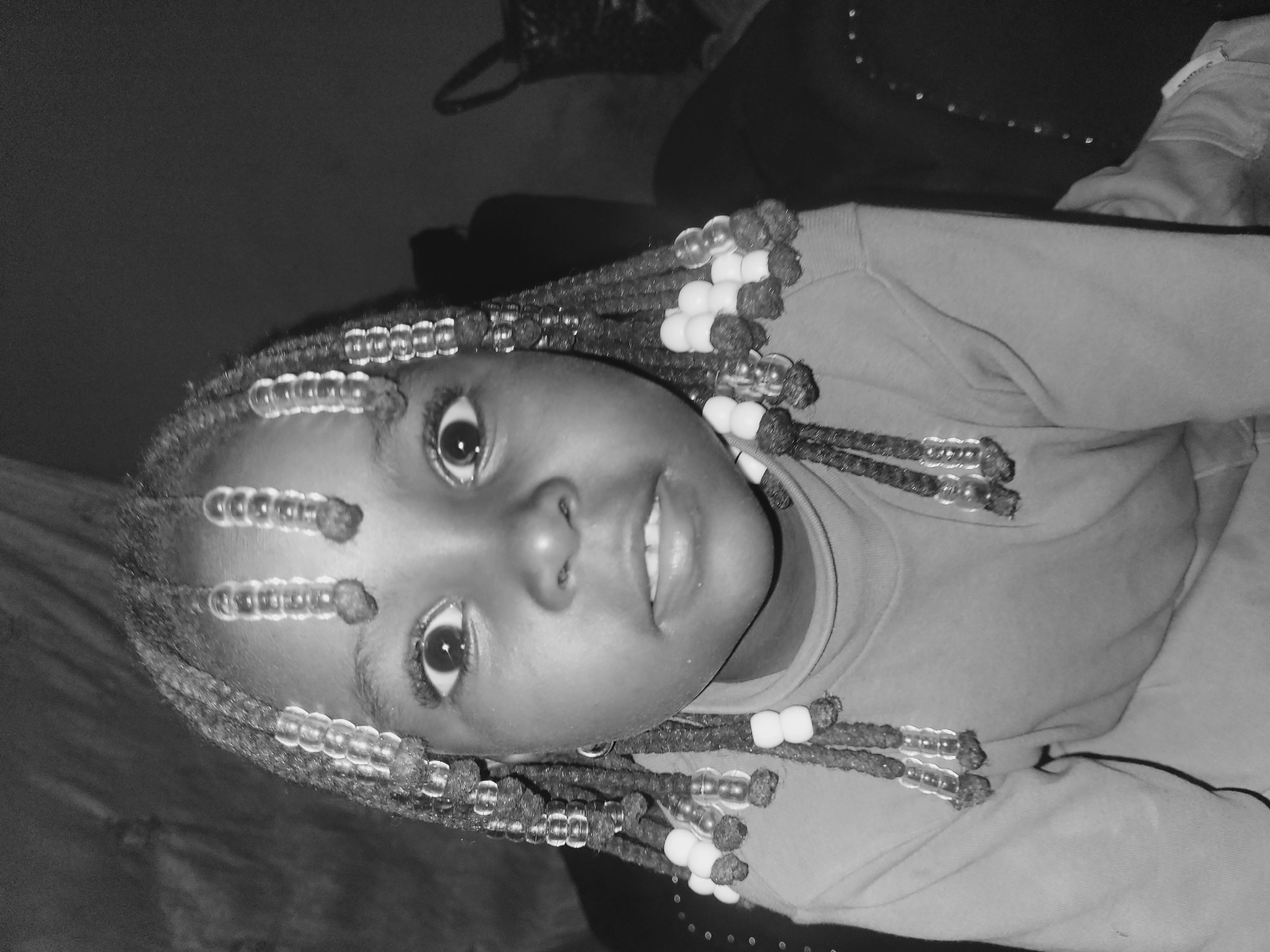 A close-up, black and white photograph shows a young, dark-skinned girl with braided hair. Her braids are adorned with beads, some clear and some white. She is wearing a t-shirt. The background is dark and out of focus, but appears to show furniture. Location: Unknown, Unknown.