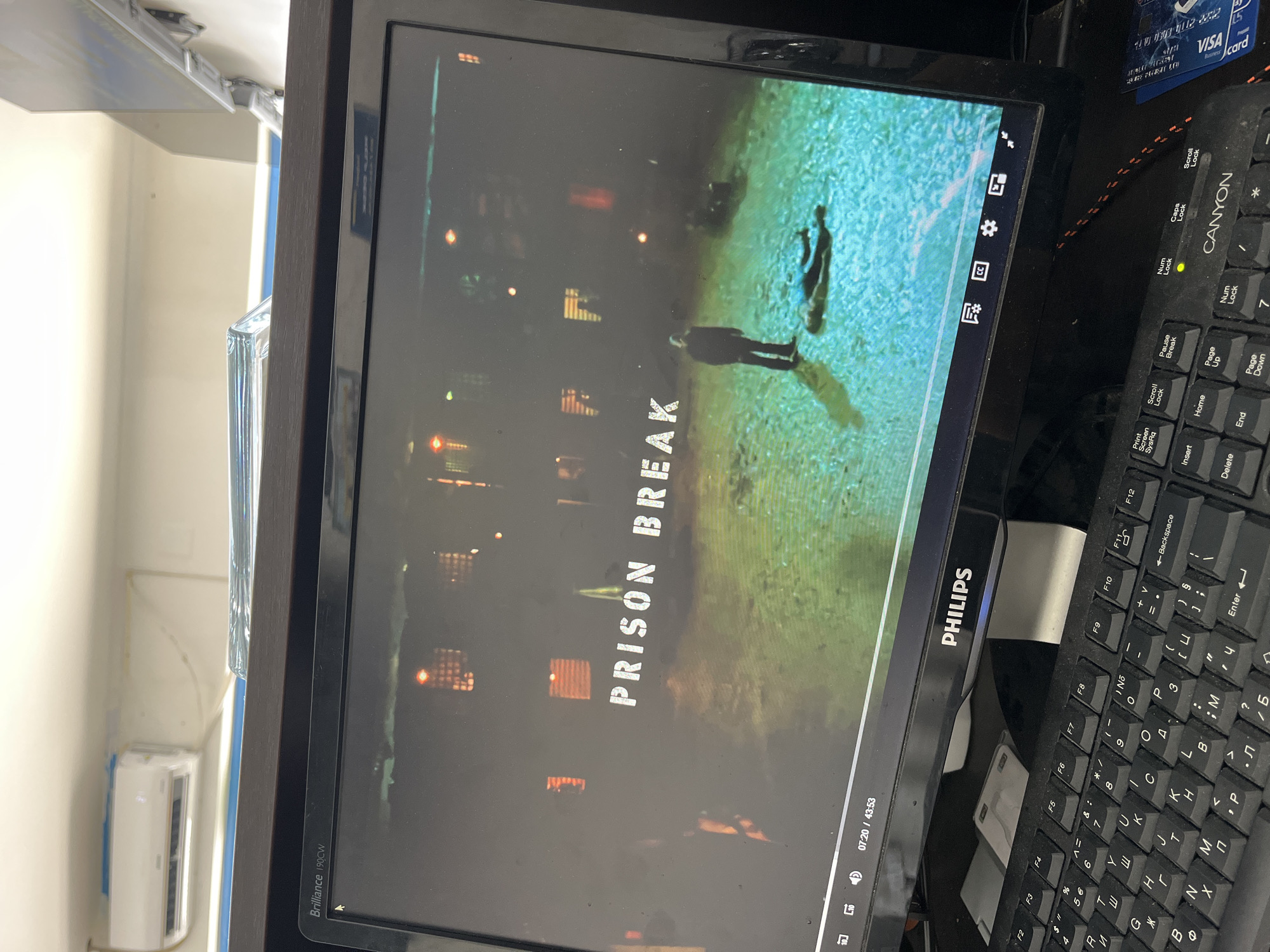 The image displays an indoor computer setup in Ivailo, Bulgaria, centered on a monitor displaying a video. A black Philips "Brilliance 190CW" monitor is prominently featured, showing content from the TV series "Prison Break." The title "PRISON BREAK" is visible on the screen, along with two silhouetted figures standing on a textured, light blue-green surface. Dimly lit shapes resembling building windows are seen in the background of the screen content. The video playback time is indicated as "07:20 / 43:53."
To the right of the monitor, a black "CANYON" brand keyboard is visible, featuring both Latin and Cyrillic characters on its keys, with the "Num Lock" light illuminated. A partial view of a "VISA" "Icard" with visible numbers is positioned above the keyboard.
The background consists of a light-colored wall with an air conditioning unit mounted on it to the left of the monitor. Overhead ceiling lights are also visible. A clear, possibly glass, object stands between the monitor and the wall on the left. The scene depicts a casual indoor setting for media consumption.