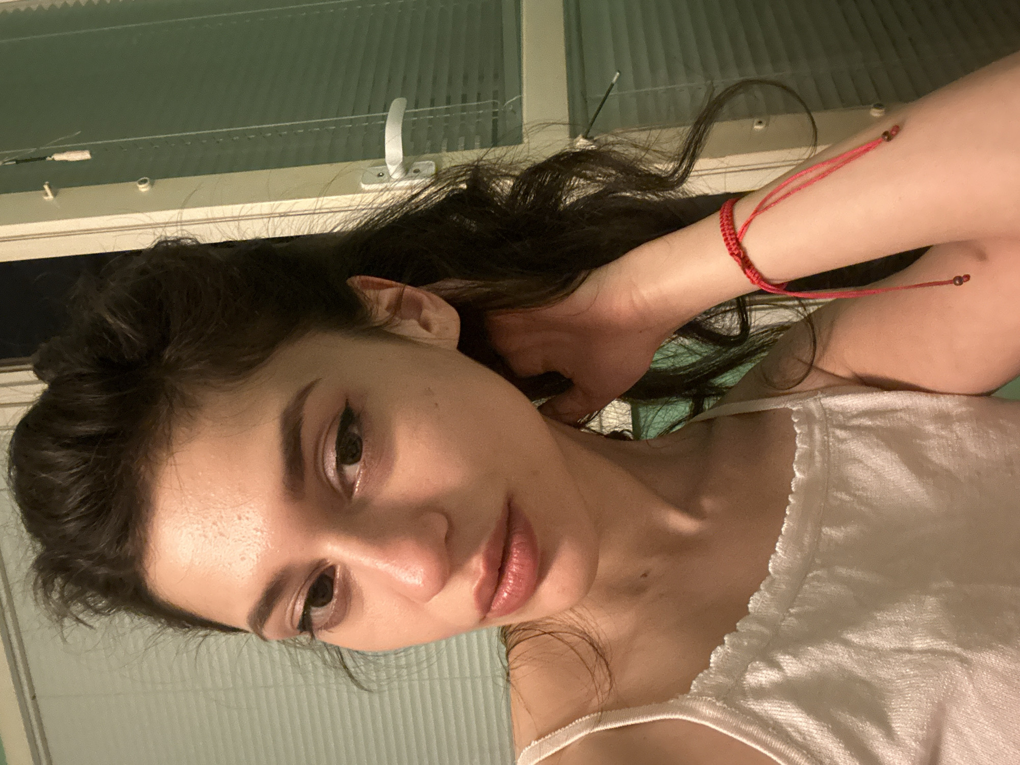 A young woman with long, dark, wavy hair, pulled back partially, is looking at the camera. She is wearing a white tank top with a scalloped neckline. She has on eyeliner and lip gloss. Her right arm is bent, and her hand is resting near her neck, revealing a red string bracelet with two small brown beads. In the background, there is a window with horizontal blinds, and a door handle on the right side. The scene appears to be indoors, likely in a residential setting. There are no visible cues to indicate a specific location in Sofia, Bulgaria.