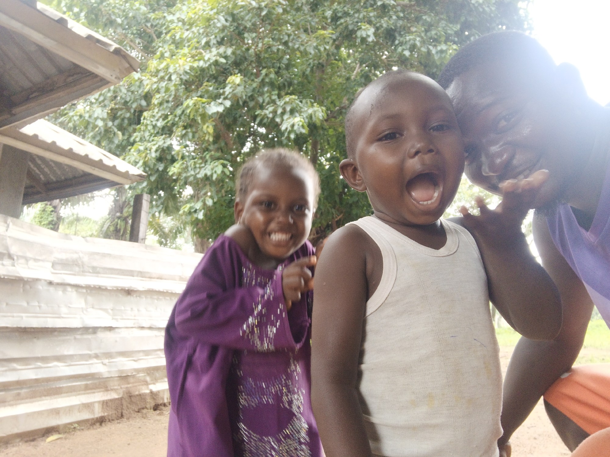 Two young children and an adult are visible in this outdoor, close-up, waist-up photograph. The child in the foreground, facing the camera, is a Black male toddler wearing a white tank top. He has an open mouth and is looking directly at the viewer. To his left, another Black child, a girl, is visible, also looking towards the camera and smiling. She is wearing a purple dress with a sparkly pattern. Partially visible on the right side of the frame is the face of a Black adult, who appears to be holding the toddler. The background consists of lush green foliage and a light-colored, corrugated metal structure that may be a wall or roof. The lighting suggests an overcast day.
