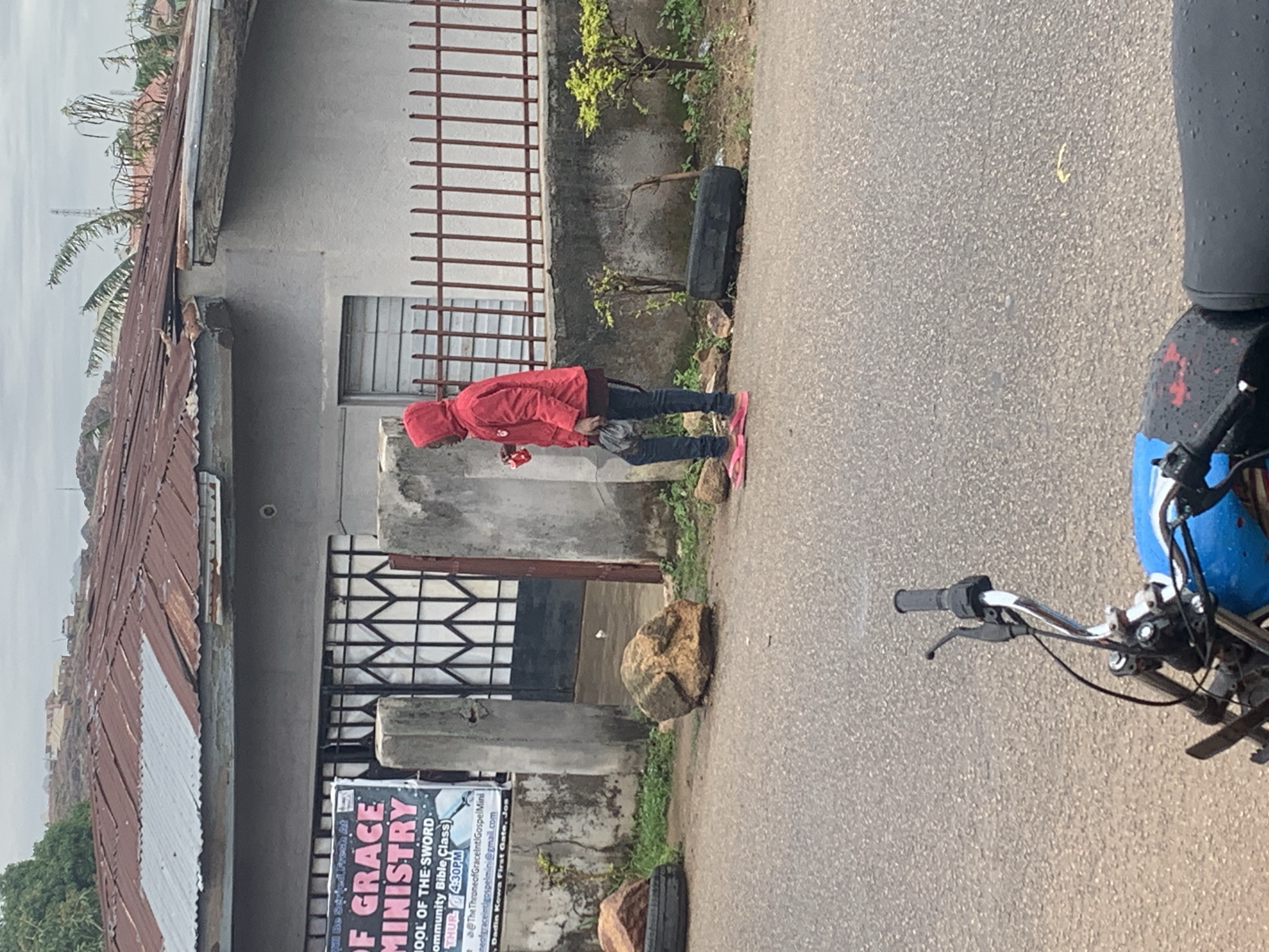 This image captures an outdoor street scene in Jos, Nigeria, showing a paved road flanked by buildings and greenery under an overcast sky. The photograph appears to be taken from a motorcycle, with a portion of its blue and black seat and handlebars visible in the right foreground.
In the mid-ground, a person wearing a red hooded jacket, dark pants, and pink flip-flops stands on the side of the road, holding a small item in their hand. Adjacent to the road, there are concrete buildings with corrugated metal roofs. Some windows feature security bars, and a metal fence is also visible. Along the roadside, there are patches of green grass and small plants, along with several rocks and a discarded black tire.
A white and black vertical banner is prominently displayed near the buildings. Visible text on the banner includes: "ill Be Surfed Fresh At OF GRACE MINISTRY SCHOOL OF THE SWORD- Community Bible Class) THUR 4:30PM @The ThroneofGraceint Gospel Mini neofgraceintigospelmini@gmail.com Dadin Kowa First Gate, Jos". This sign indicates a religious event or organization, and the mention of "Jos" corroborates the geographical context. The overall lighting suggests it is either a cloudy day or early morning/late afternoon.