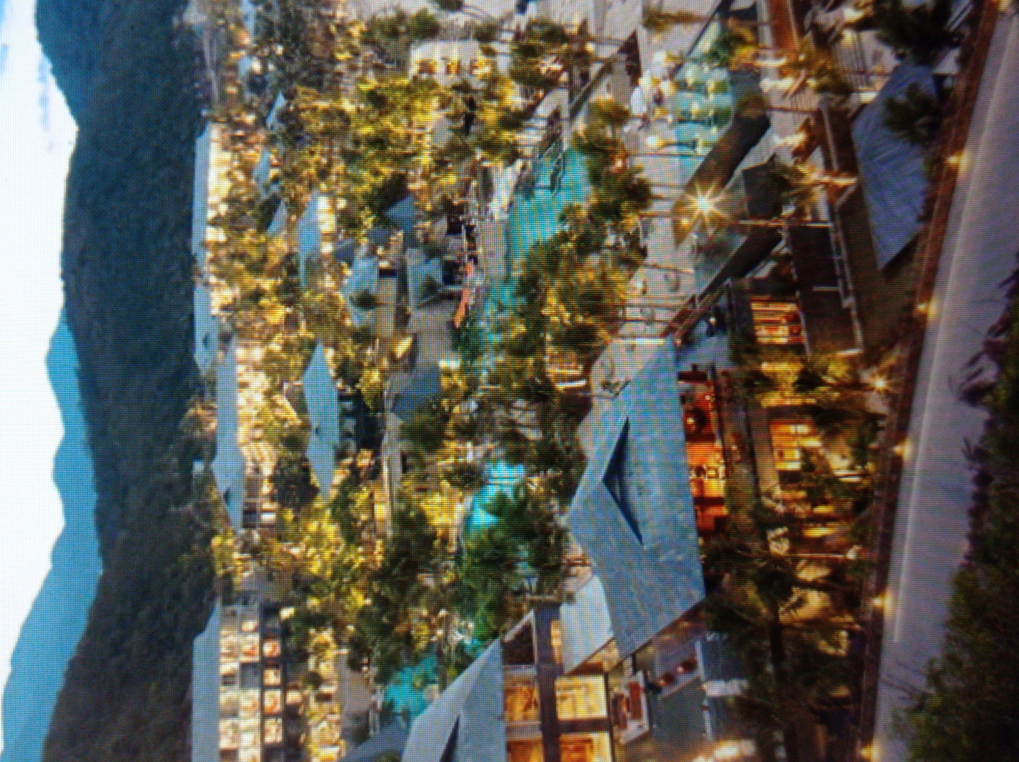 The image depicts an aerial view of a resort complex nestled against a backdrop of verdant, mountainous terrain. The resort features multiple multi-story buildings with modern architecture, characterized by flat roofs and extensive windowing. Several swimming pools are visible, interspersed with lush tropical vegetation and palm trees. The complex is illuminated, suggesting it is either dusk or nighttime, with warm lights emanating from within the buildings and along pathways. The overall impression is one of a luxurious and well-maintained leisure destination. The image does not contain any elements that would suggest it is located in the city of Mukachevo, Ukraine.