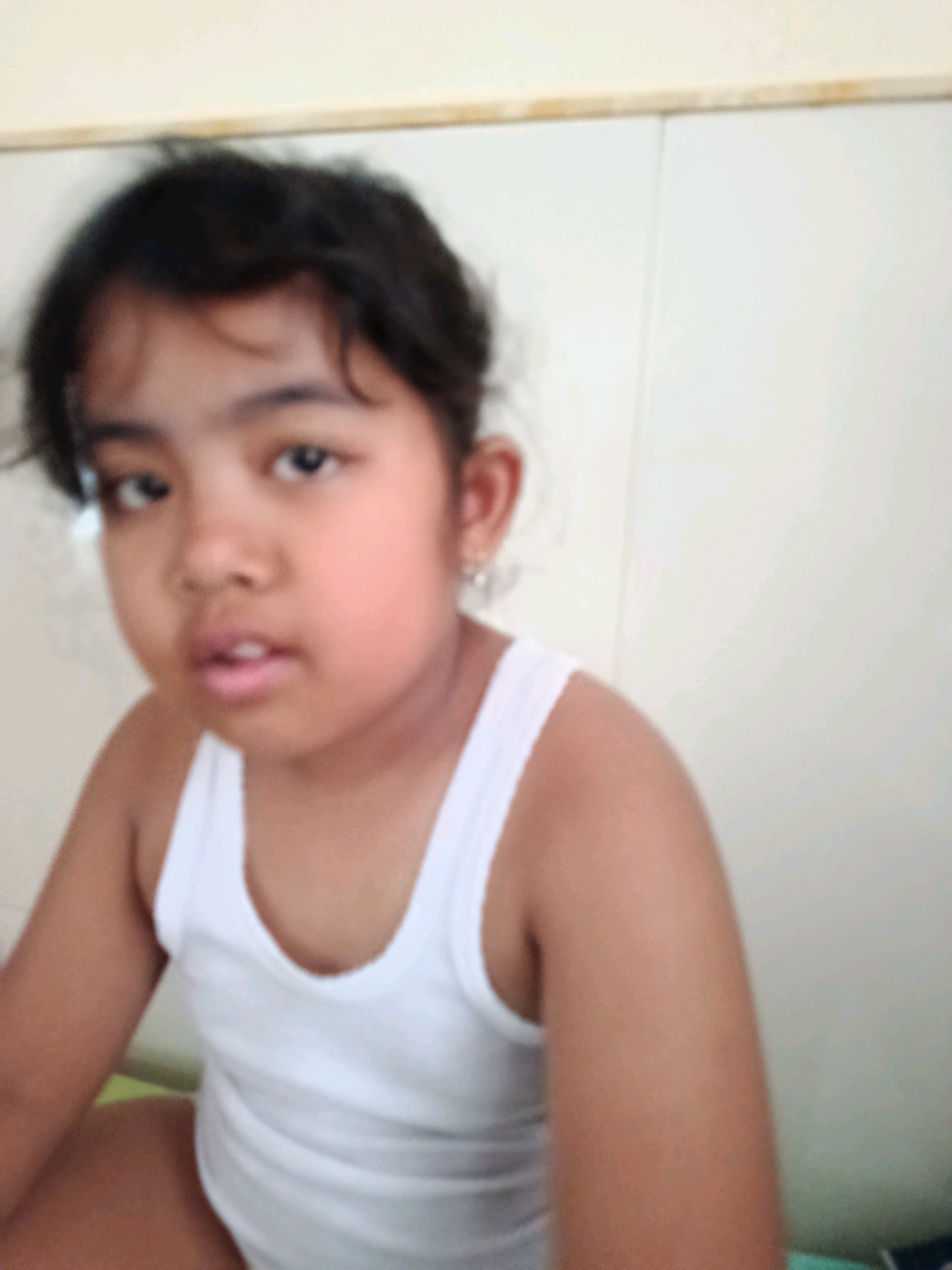 A young girl is captured in a close-up portrait, wearing a white tank top. Her dark hair is styled loosely around her face, framing her features. Her eyes, dark and expressive, gaze towards the viewer. A subtle hint of pink blush appears on her cheeks, and her mouth is slightly open, as if she is about to speak. She has a small earring in her left ear. The background is a plain, light-colored wall, with a slight shadow line running vertically down the middle, suggesting it might be two panels. The lighting is soft and diffused, with no strong shadows, hinting at an indoor setting. The overall impression is one of innocence and gentle curiosity. The time of day and weather are not discernible from the image. There is no visible text in the media file.