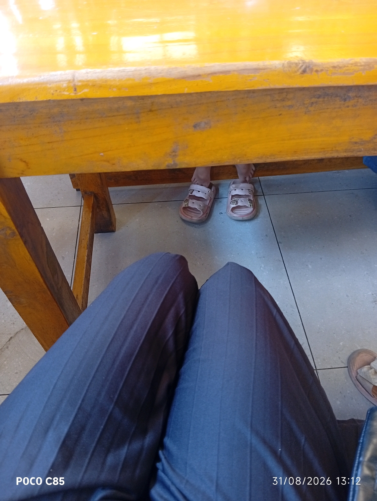 The image depicts a low-angle shot taken from beneath a yellow wooden table. On the floor, which appears to be tiled, are two pairs of children's sandals. One pair is pink with cartoon character embellishments and is worn by a child whose feet are visible. The other pair of sandals is visible but not on a person's feet. In the foreground, a person's legs are visible, clad in dark, pleated pants. The lower left corner of the image shows "POCO C85" and the lower right corner displays "31/08/2026 13:12". The overall environment suggests an indoor setting, possibly a classroom or dining area. The city of South Tangerang, Indonesia, is not explicitly identifiable from the visual cues in the image.