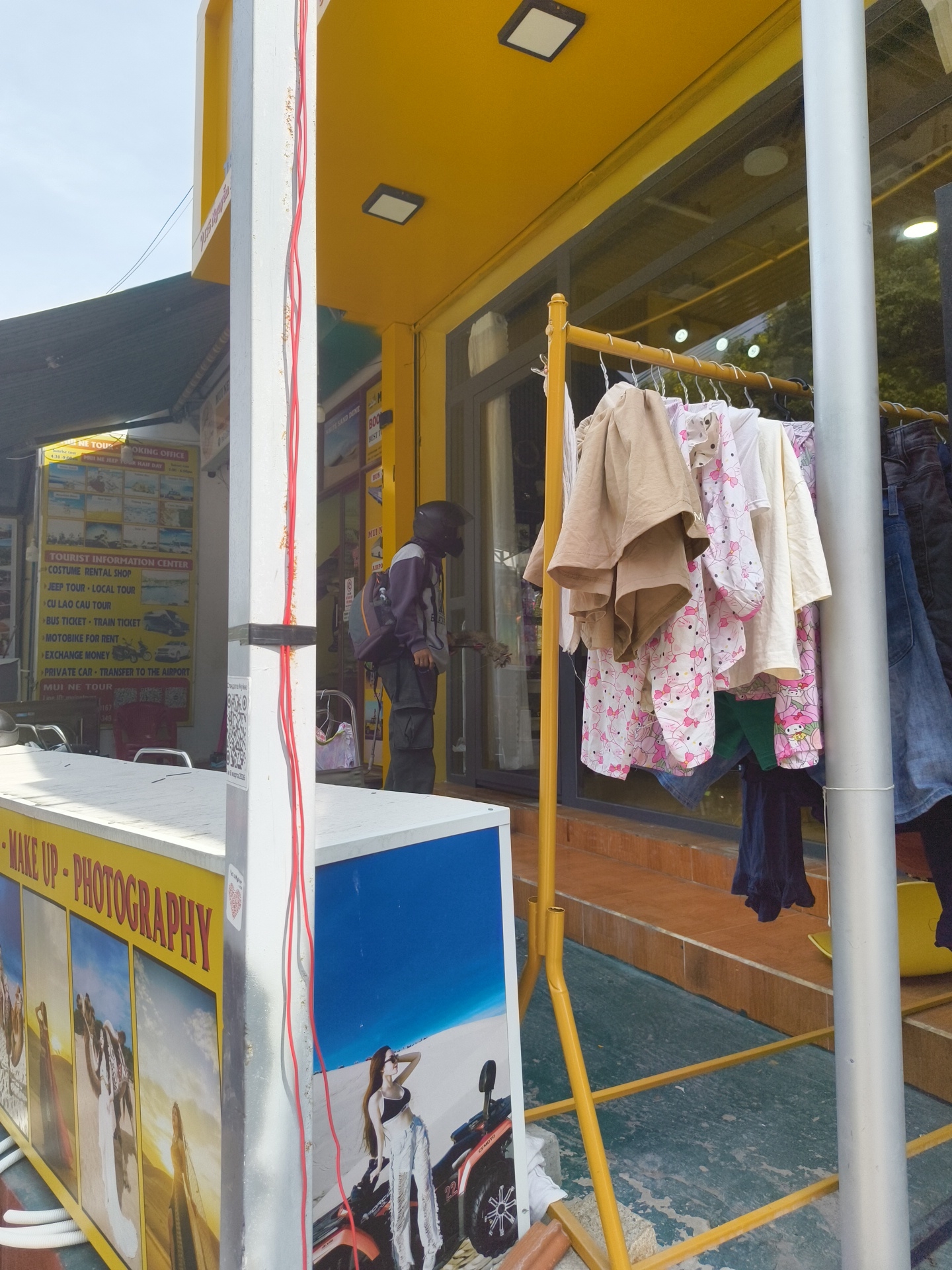 The image depicts an outdoor scene in Phu Thuy Ward, Vietnam. A person wearing a dark helmet and a backpack stands near the entrance of a building with a bright yellow facade. In front of the building, clothes are hanging on a yellow clothes rack. To the left of the building, a white structure with yellow signage displays images and text related to "MAKE UP - PHOTOGRAPHY." The ground is a mottled blue and green surface, and some stairs are visible in the background. A white pole with red wires running along it is prominently positioned in the foreground.