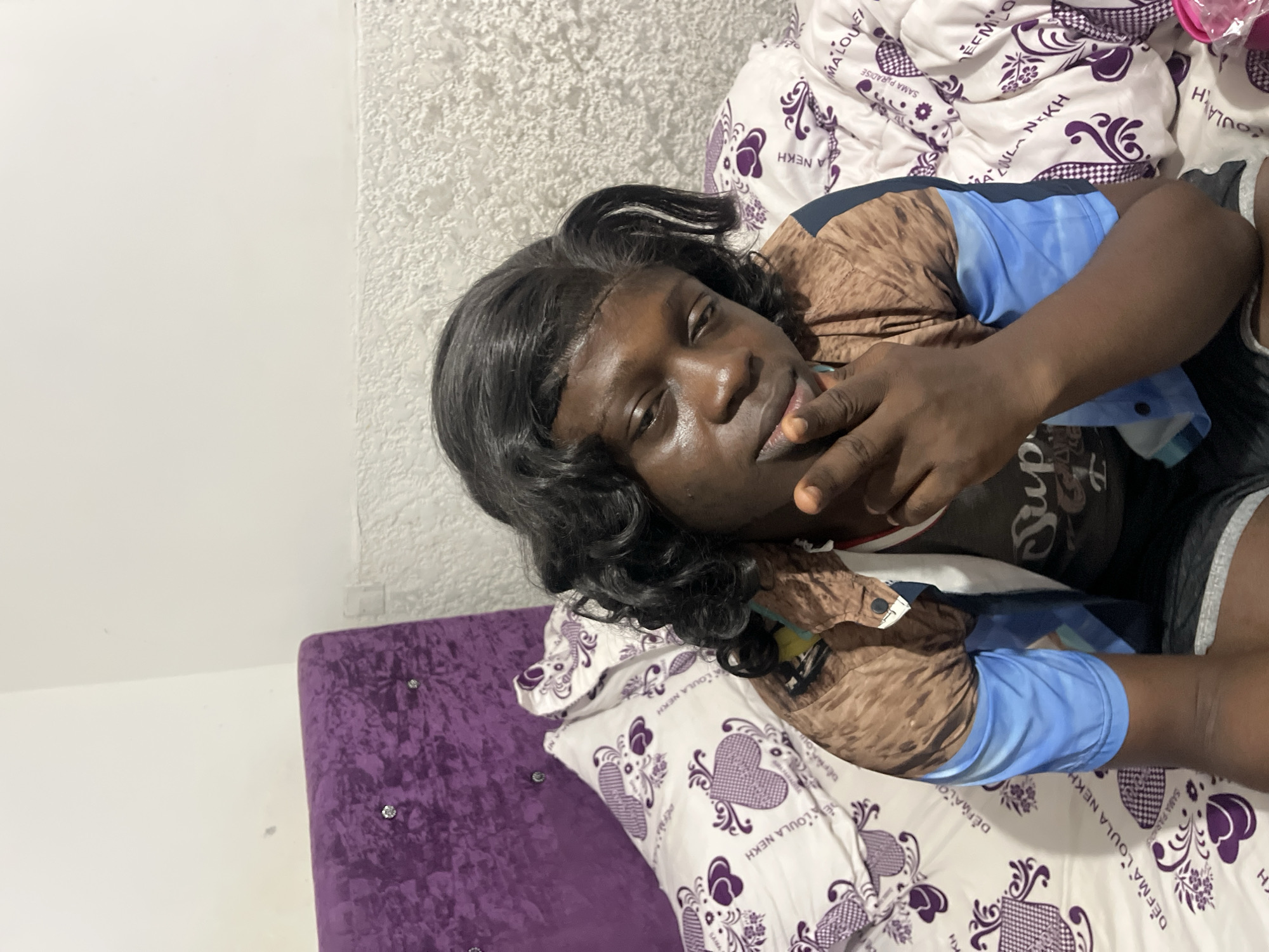 A dark-skinned person with dark curly hair (possibly a wig) is lying on a bed or lounge. The person is wearing a patterned brown and blue shirt and shorts. The bed has a white pillow and comforter with purple heart and floral patterns, with "DEFMA LOULA NEKH" and "SAMA PARADISE" written on them. A purple, button-tufted headboard or side table is visible near the person's head. The background wall is white with a textured pattern. The image does not provide sufficient details to determine the location in Abuja, Nigeria.