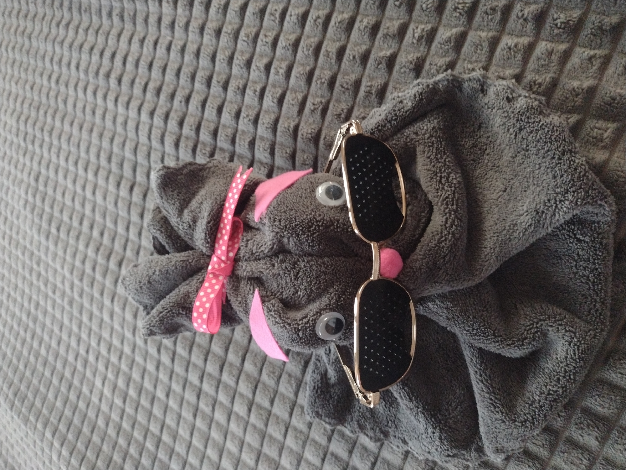 The image presents a still life arrangement on a gray, textured surface. A gray towel is folded and styled to resemble an animal, with pink felt ears and a pink ribbon. Two googly eyes are attached, and a pair of sunglasses with gold frames is placed on top of the towel. The location is Burgas, Bulgaria.