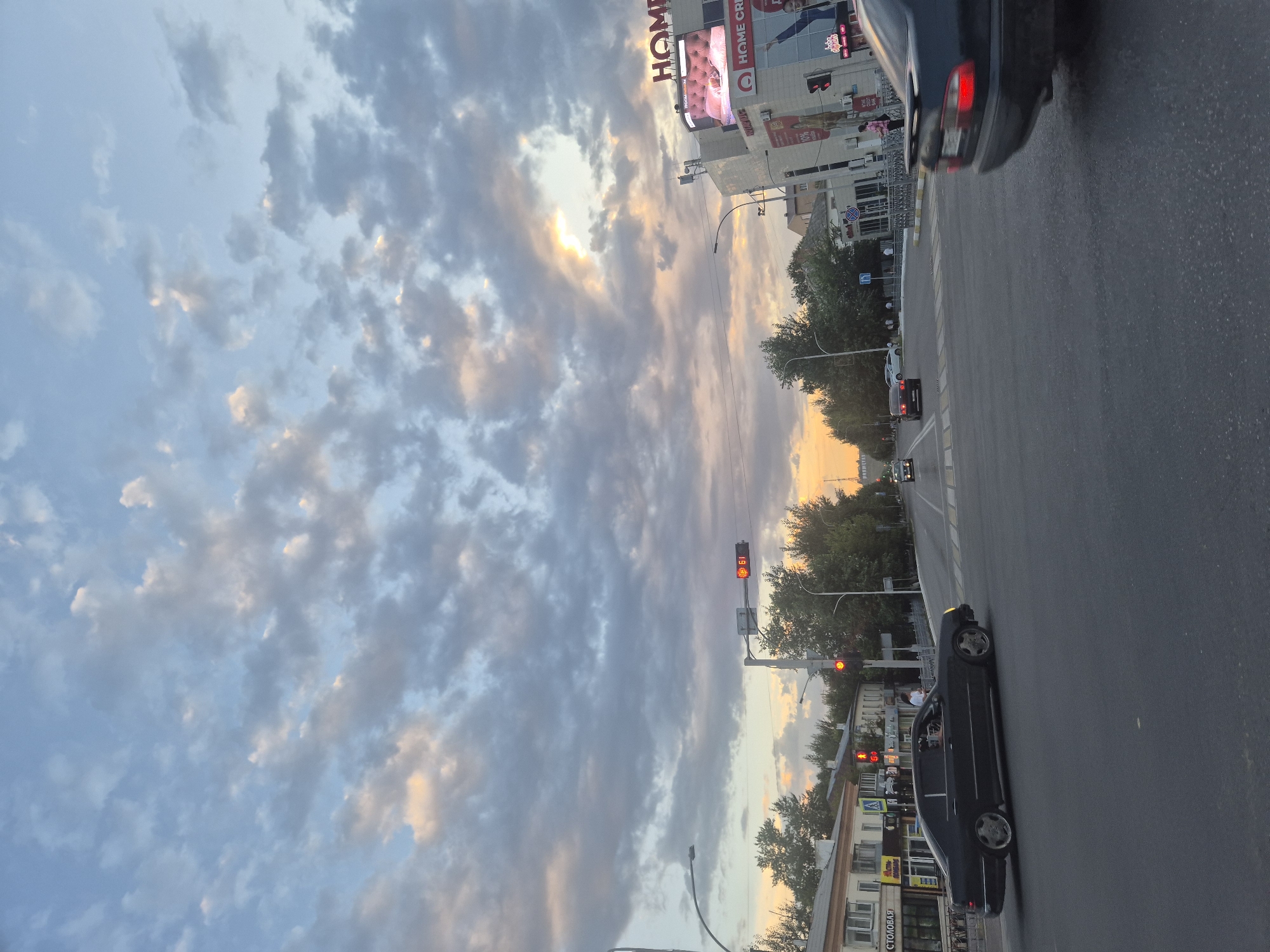 The image depicts a city street intersection in Kokshetau, Kazakhstan, during the golden hour. The sky is filled with dramatic, scattered clouds, with a bright patch of sunlight peeking through near the horizon. On the right side of the street, a multi-story building displays a large "HOME" sign and advertising screens, suggesting a commercial establishment. Traffic lights are visible, with red lights illuminated, indicating traffic is stopped. Several cars are present, including a dark sedan in the foreground and other vehicles in the background. Pedestrians can be seen on the sidewalks and crossing the street. Trees line the sides of the road, and buildings with shops are visible on the left. The overall scene captures a moment of urban life at dusk, with the warm glow of the setting sun illuminating the cityscape.