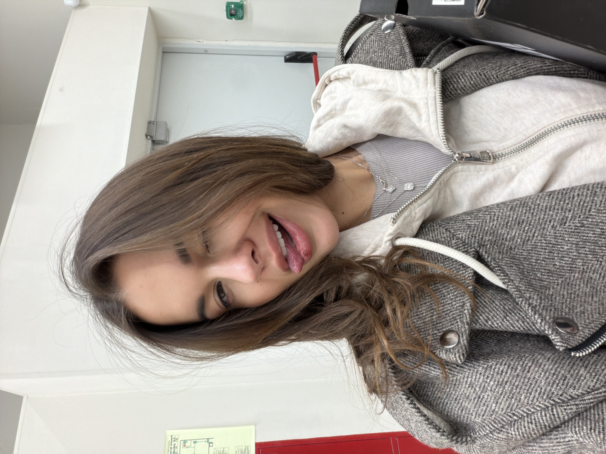 A young woman is captured in a playful indoor selfie, situated in Krasnoyarsk, Russia. She is shown from the chest up, with her head tilted, playfully winking with her right eye and sticking her tongue out. Her expression is lighthearted and candid.
She has light brown to auburn hair that falls around her face and shoulders. Her attire consists of a light grey ribbed top, layered with a light cream-colored zippered hoodie, and an outer grey jacket with a distinctive herringbone pattern. This jacket features large metallic snap buttons and a visible zipper. She wears two delicate silver necklaces, each adorned with small pendants, resting on her collarbone.
The background indicates an indoor setting, likely a hallway or a public building. The walls are predominantly white, and a white door with a silver handle mechanism is visible on the right. Above the door, a small green square object is mounted on the wall. In the lower left corner, a white-framed diagram or chart, possibly a floor plan or safety instruction, is affixed to the wall, with unreadable text and colored sections. Below this, a solid red panel or surface can be seen. The lighting is bright and even, typical of interior artificial light. No legible text is visible in the image.