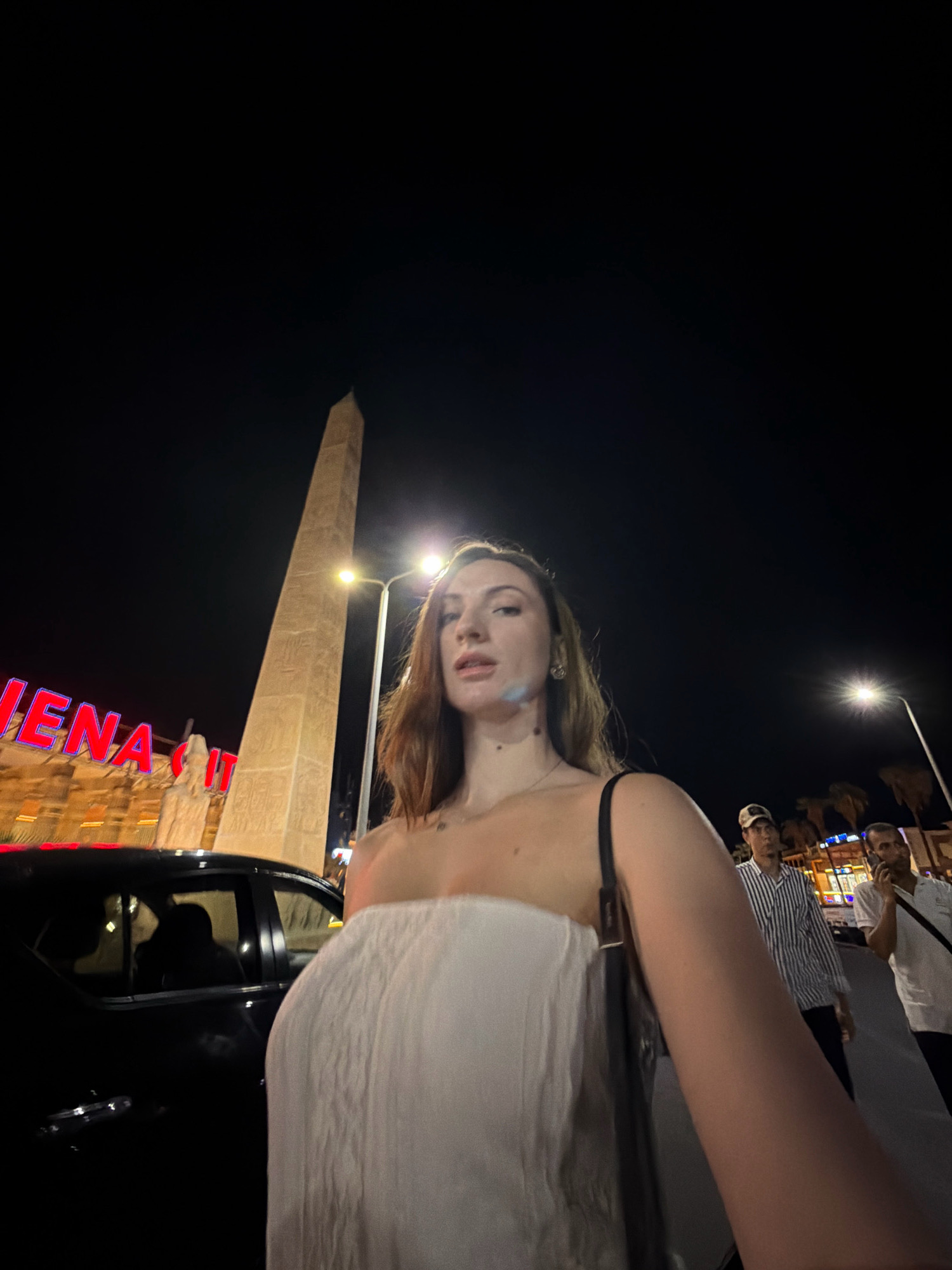 An outdoor, nighttime, wide-angle, selfie-style photograph shows a Caucasian woman in the foreground, facing the camera. She has brown hair and is wearing a strapless white garment. She is standing next to a black car. In the background, an obelisk is visible, with hieroglyphics etched into its surface. To the left of the obelisk, a neon sign reads "VENA CITY" in blue and red letters. Several people are walking in the background, and palm trees are visible. The scene appears to be in Sharm El Sheikh, Egypt, a popular tourist destination known for its ancient Egyptian-themed attractions.