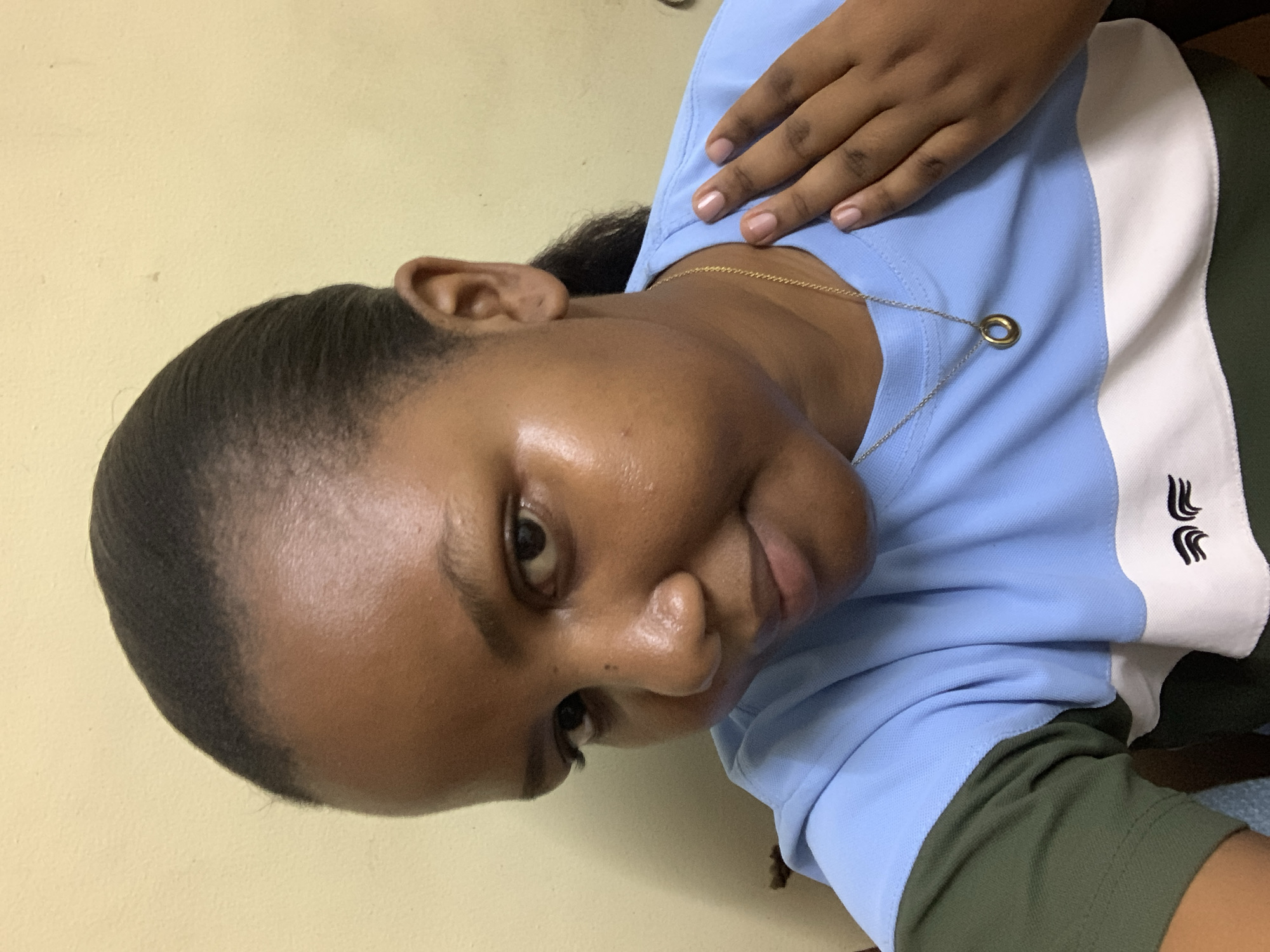 This close-up, vertical selfie features a person, likely female, with medium-dark skin and dark hair pulled back. The subject's head is tilted slightly, and they look towards the camera with a subtle, neutral expression. Highlights are visible on the forehead and nose, indicating smooth skin.
The individual is dressed in a collared polo shirt, which is light blue on the left side, transitioning to a vertical white stripe, and then a dark green section on the right. A small black embroidered logo, resembling three stylized wave-like symbols, is visible on the white stripe of the shirt. A delicate gold-colored chain necklace with a small, circular gold pendant rests on the light blue fabric. One hand, adorned with light-colored manicured nails, is gently placed on the upper chest area of the shirt.
The background is a plain, light yellow or beige wall, suggesting an indoor environment. The lighting is even, typical of indoor illumination. The image captures a casual moment in Dar es-Salaam, Tanzania. No specific time of day or weather conditions can be determined from the frame.
