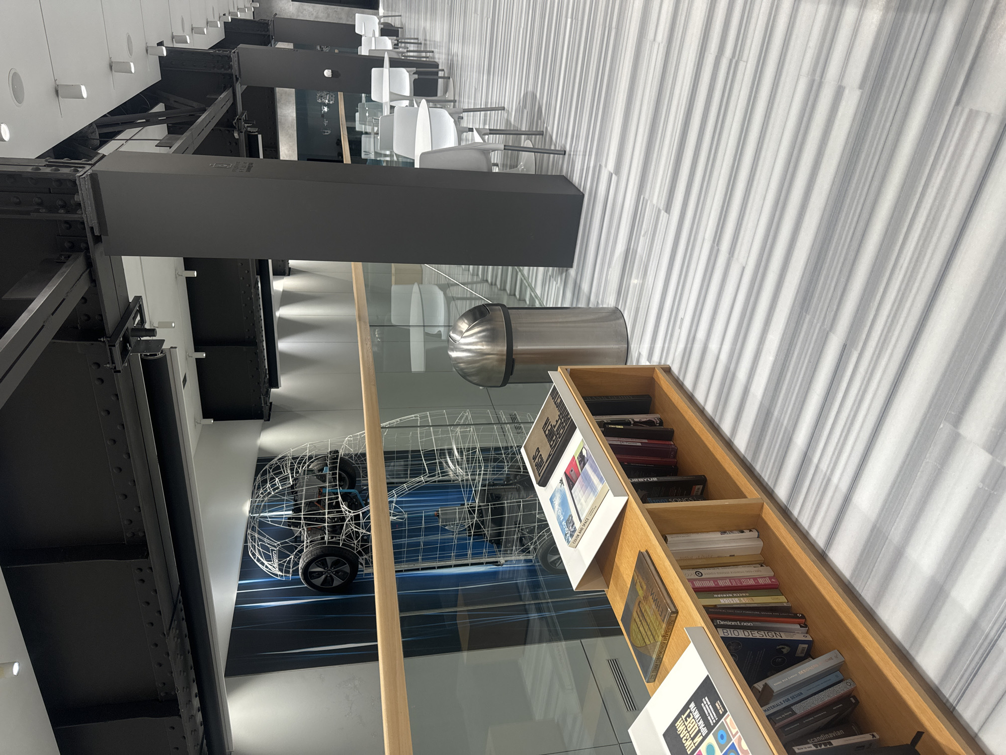 The image shows an interior space with modern architectural elements. A prominent feature is a metal framework, possibly part of the building's structural design, painted in dark grey or black. There are also rows of chairs and tables, suggesting a cafe or lounge area. A bookshelf filled with books is visible, and there's a metallic trash can. A wireframe model of a car is displayed, hinting at a design or engineering context. The environment suggests a contemporary, possibly industrial-chic, space.
The OCR labels on the books on the bookshelf indicate the design, art, and architecture theme of the place. Some recognizable words are "DESIGN," "COLOR AND SPACE," "ARCHITECTURE" and "MATERIALS FOR DESIGN."
Due to the sparse information available, it's impossible to conclusively determine the exact location. If there is more information available, the exact location in Moscow might be provided.