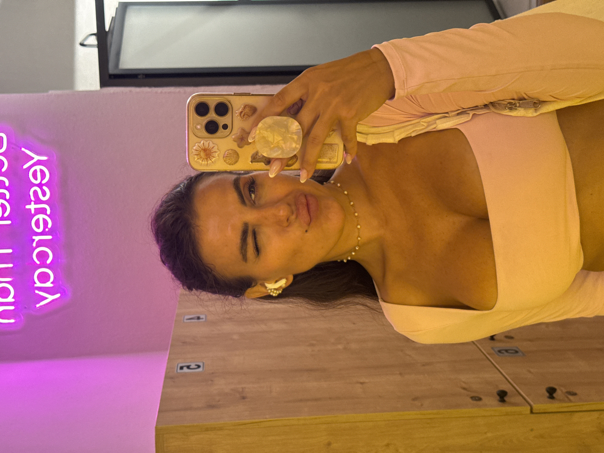 The image captures a woman taking a selfie in a locker room. She is wearing a light pink top and has a pearl necklace. Her phone, which she is holding up to take the photo, has a gold case adorned with seashells and a circular grip. A neon sign, which reads "Lucky Yesterday" in reverse, is visible on the purple wall behind her. The locker room has wooden lockers with numbers on them, suggesting a gym or a similar facility. The lighting appears to be artificial and warm, casting a yellow glow on the right side of the image. The woman is winking and making a playful expression. The time of day is not discernible, but the indoor setting suggests it could be any time. The scene appears to be set in Fethiye, Turkey.