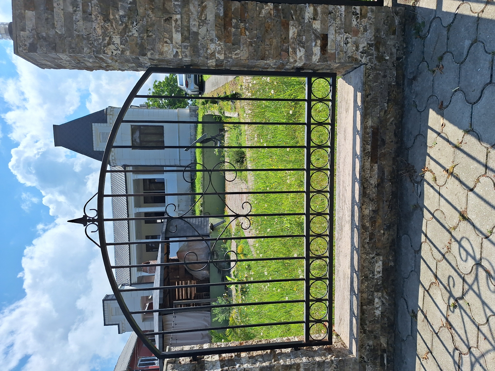 The image shows a wrought iron gate in front of a residential property in Kobuleti, Georgia. The gate is black and features decorative scrollwork and circular elements. It is flanked by stone pillars. Behind the gate, a lawn with scattered white flowers leads to a white house with columns and a dark, conical roof. A white car is parked on a driveway. The sky is blue with fluffy white clouds. The foreground shows a patterned paved area with shadows cast by the gate.