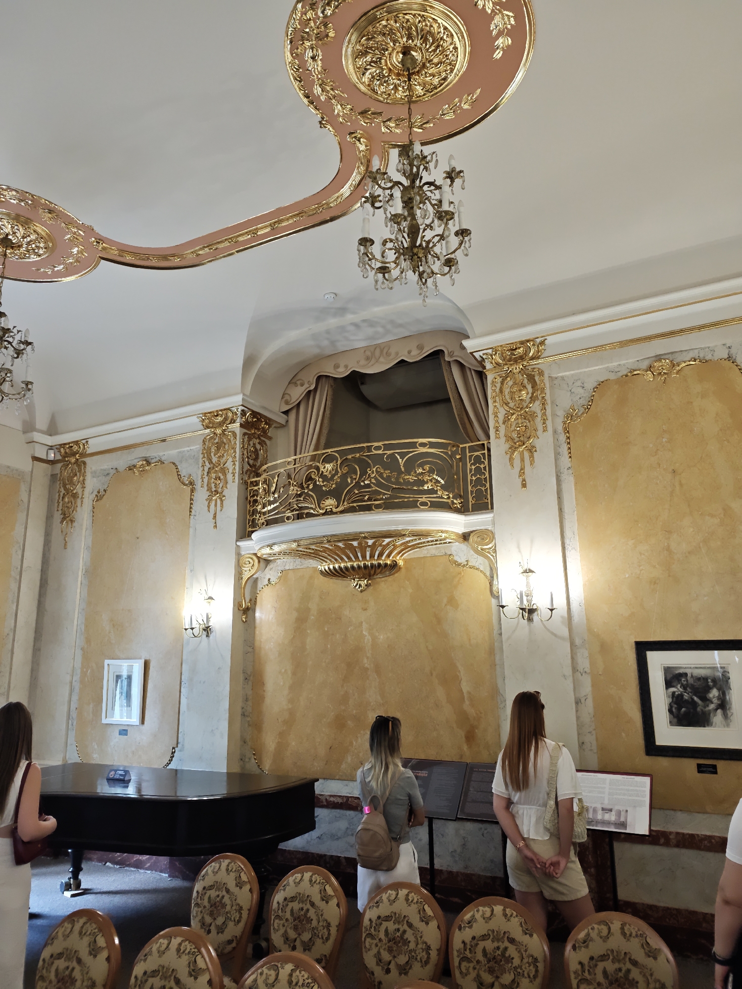 The image depicts the interior of a grand, opulent room, likely a museum or historic building, with ornate gold embellishments and a light pink and gold ceiling. In the foreground, a grand piano is visible, and several chairs with patterned upholstery are arranged in front of it. Three women are present, facing away from the camera, observing exhibits or the room's features. One woman on the left is wearing a white dress and carrying a red bag. Another woman in the center wears a grey top and a backpack, and the woman on the right is in a white top and beige shorts. The room has yellow marble-patterned walls, a balcony with gold railings, and chandeliers hanging from the ceiling. On the wall, there is a framed picture and informational displays. The style suggests it could be a preserved historical space within Lviv, Ukraine, potentially related to music or a significant cultural figure, given the presence of the piano.