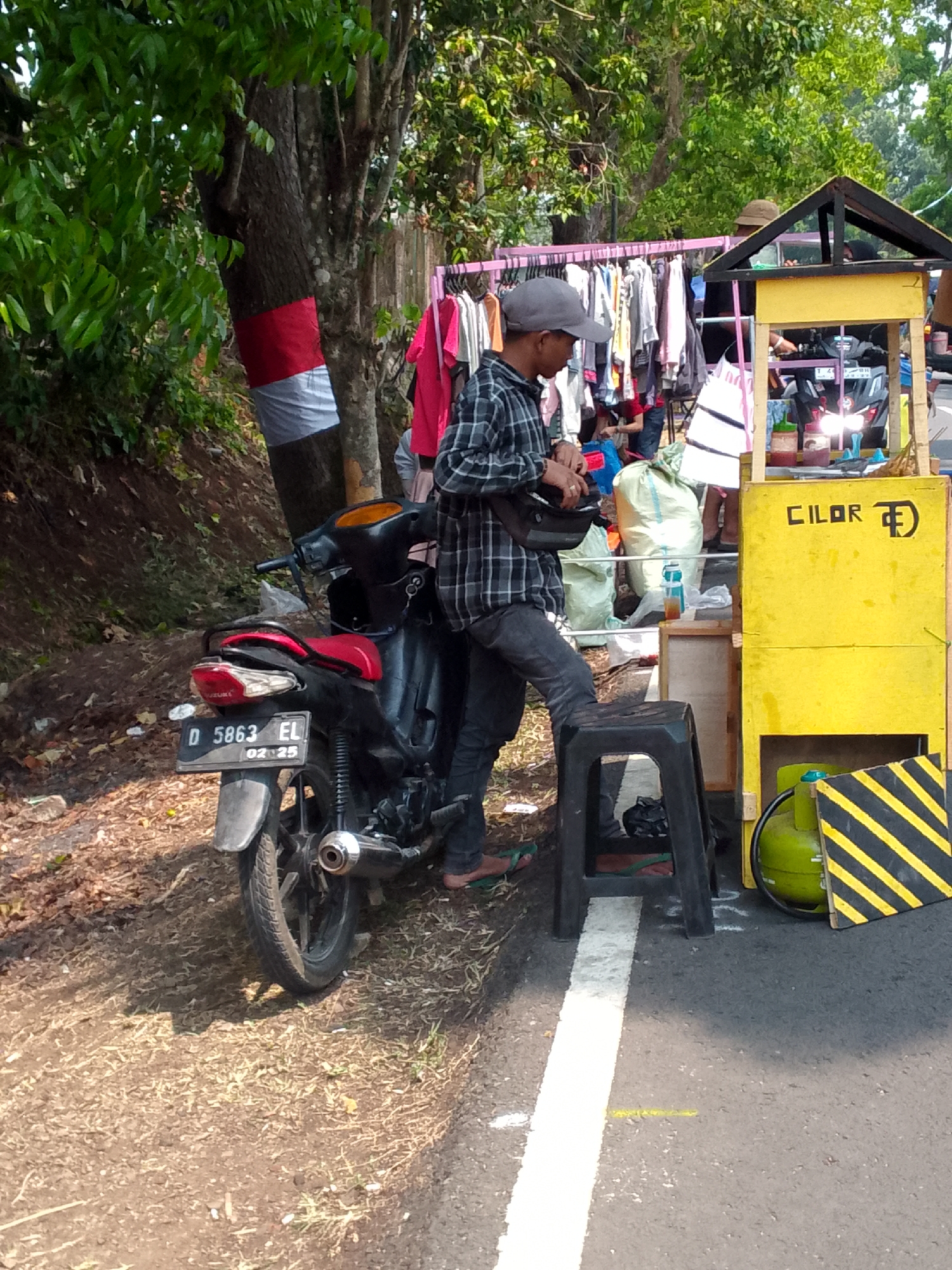The image depicts a street scene in Mandalawangi, Indonesia, likely a roadside vendor setup. A young man wearing a plaid shirt and a grey cap stands next to a parked black motorcycle with a red seat. He is holding a black bag and appears to be engaged in a transaction or preparing to serve a customer.
To his right is a small, brightly colored yellow stand with a wooden roof. The stand has the word "CILOR" written on it in black letters, along with a logo. Inside the stand, there are bottles of sauces, a gas cylinder, and other items associated with food vending. A rack of clothes is visible in the background, suggesting a multi-purpose vendor.
The environment is outdoors, with a dirt and grass embankment to the left and a paved road with a white line to the right. Trees with green foliage provide shade. The overall impression is of a typical bustling street market in a tropical climate.