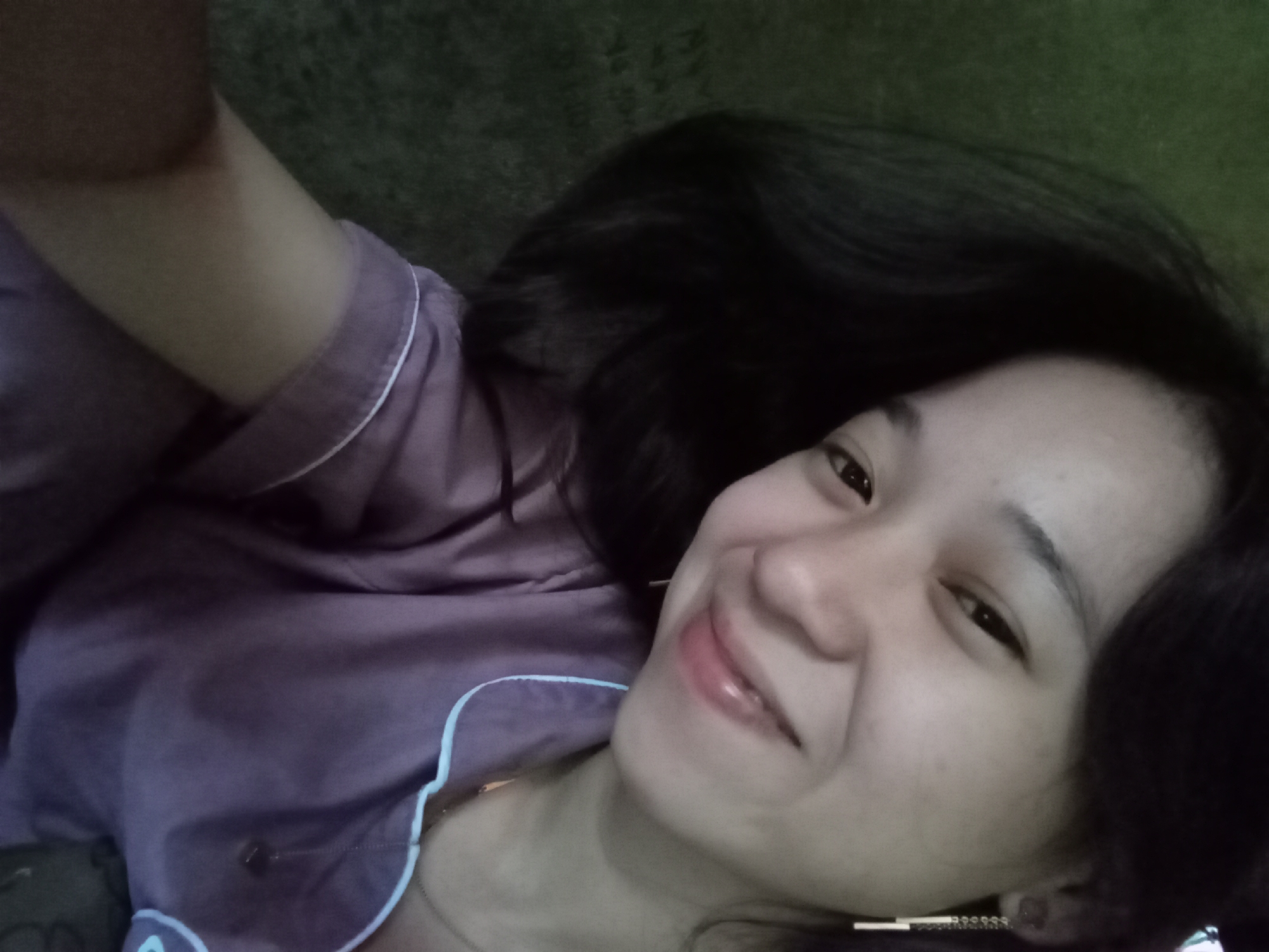 A young woman with dark hair, wearing purple pajamas, smiles directly at the camera. She is wearing a long, dangling earring on her right ear. Her left arm is bent at the elbow and extended towards the camera. The background is a dark, textured surface, possibly a wall or fabric. The lighting is dim. The image appears to have been taken indoors, likely in a residential setting. No specific location in Rejosari, Indonesia can be determined from the visual information provided.