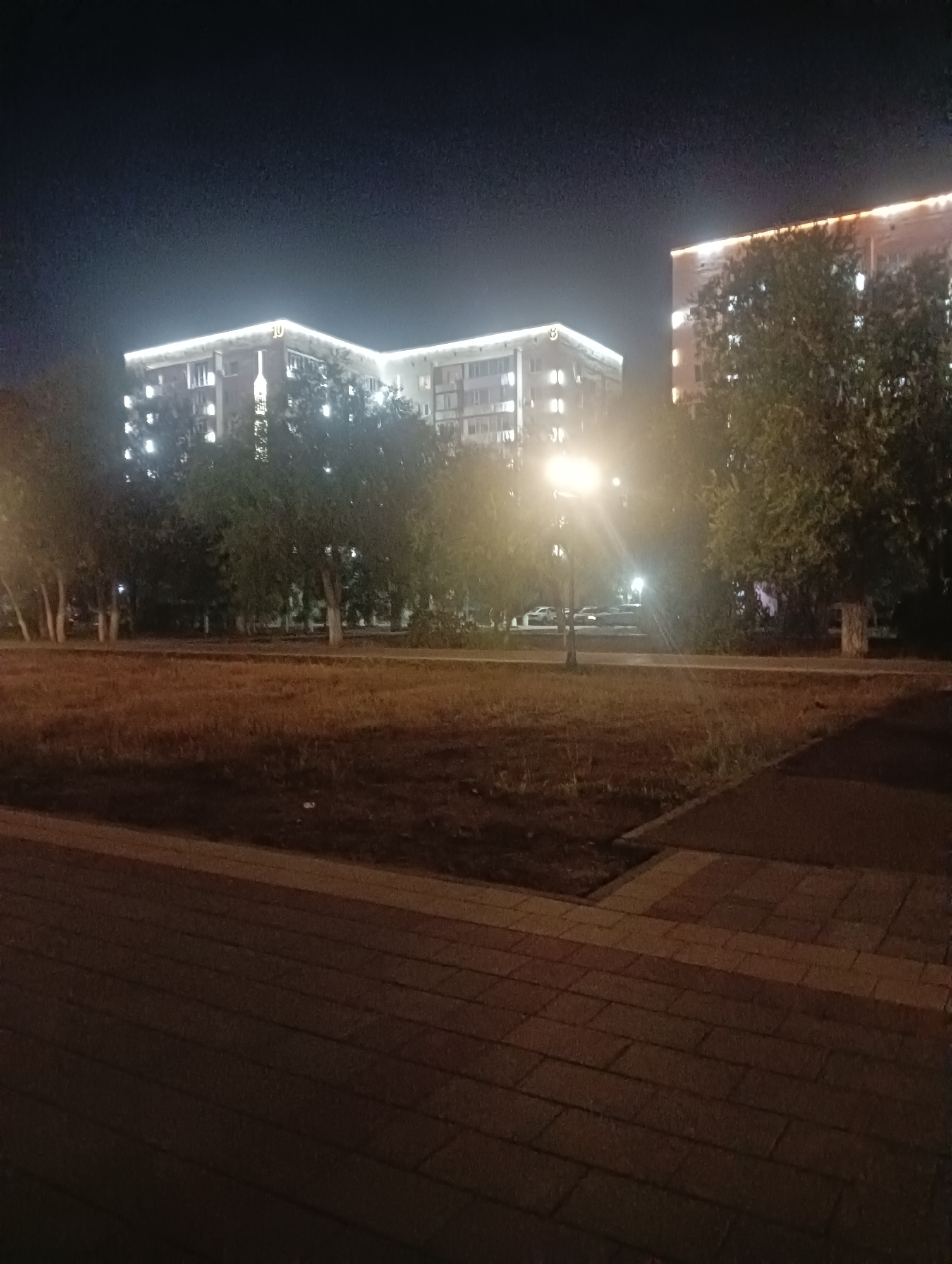 The image captures a nighttime scene in the city of Aktobe, Kazakhstan. Two multi-story apartment buildings are visible, illuminated by exterior lighting that outlines their structures. The building on the left has the Cyrillic letter "Ю" prominently displayed on its facade. Trees, some with bare branches and others with foliage, are scattered in the foreground and between the buildings, partially obscuring them. A streetlamp with a bright light casts a glow on the surrounding area. The ground in the foreground is paved with brick-like tiles, and a patch of dry grass or soil is visible between the pavement and the buildings. The overall atmosphere is calm and quiet, typical of a residential area at night.
