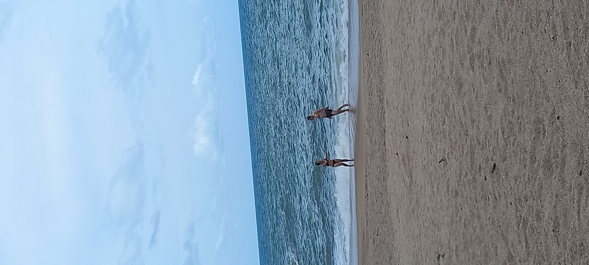 The image depicts a beach scene in Baan Thong Nang, Thailand. The sky is partly cloudy. The ocean occupies the central part of the image, with waves lapping onto the sandy beach. Two people are standing near the water's edge. One appears to be a man, wearing dark-colored swimwear. The other person appears to be a woman, also in swimwear. They appear to be wading in the water.